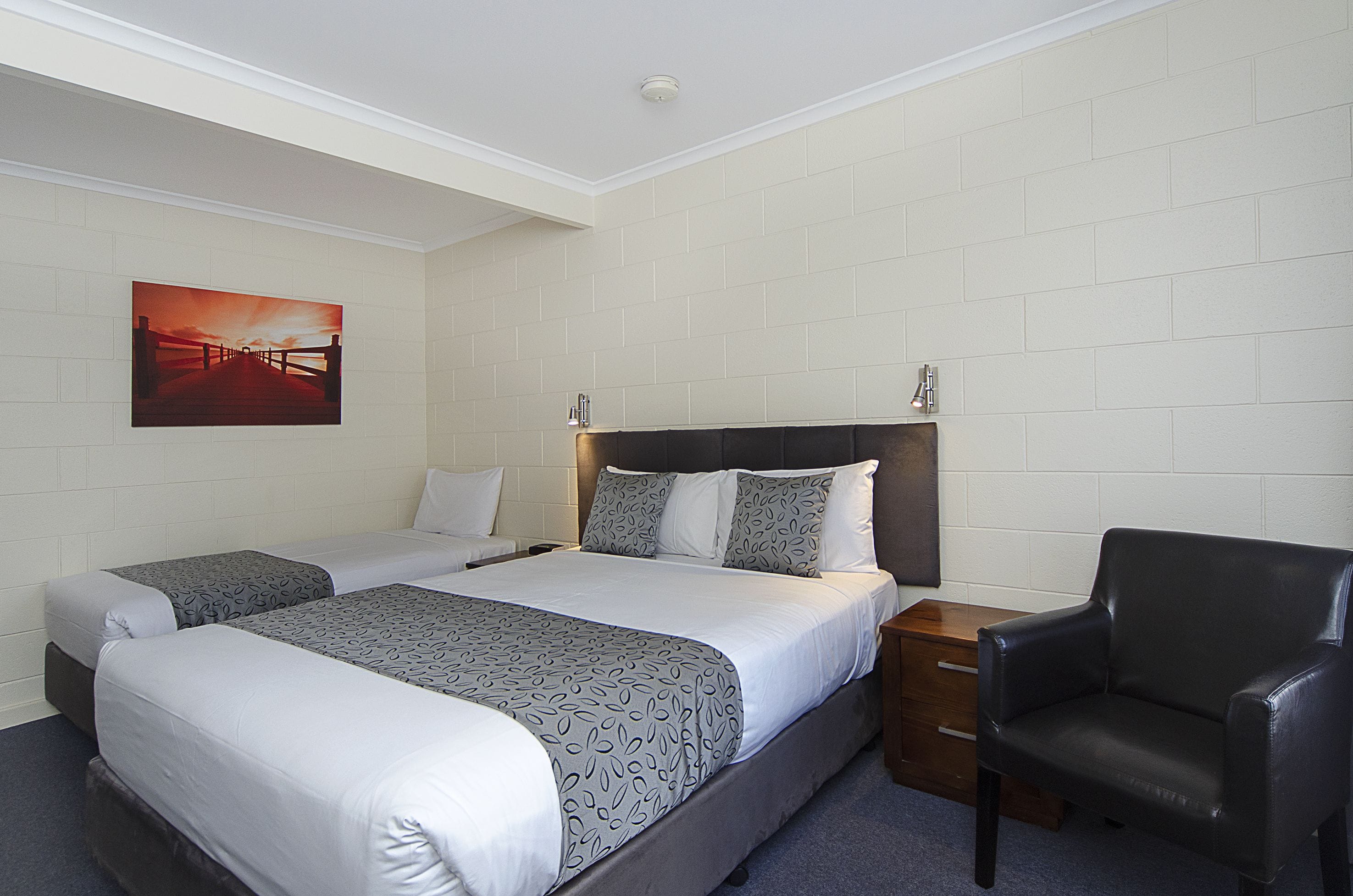 Habitación de hotel con dos camas, una mesita de noche, una silla y un cuadro en la pared.