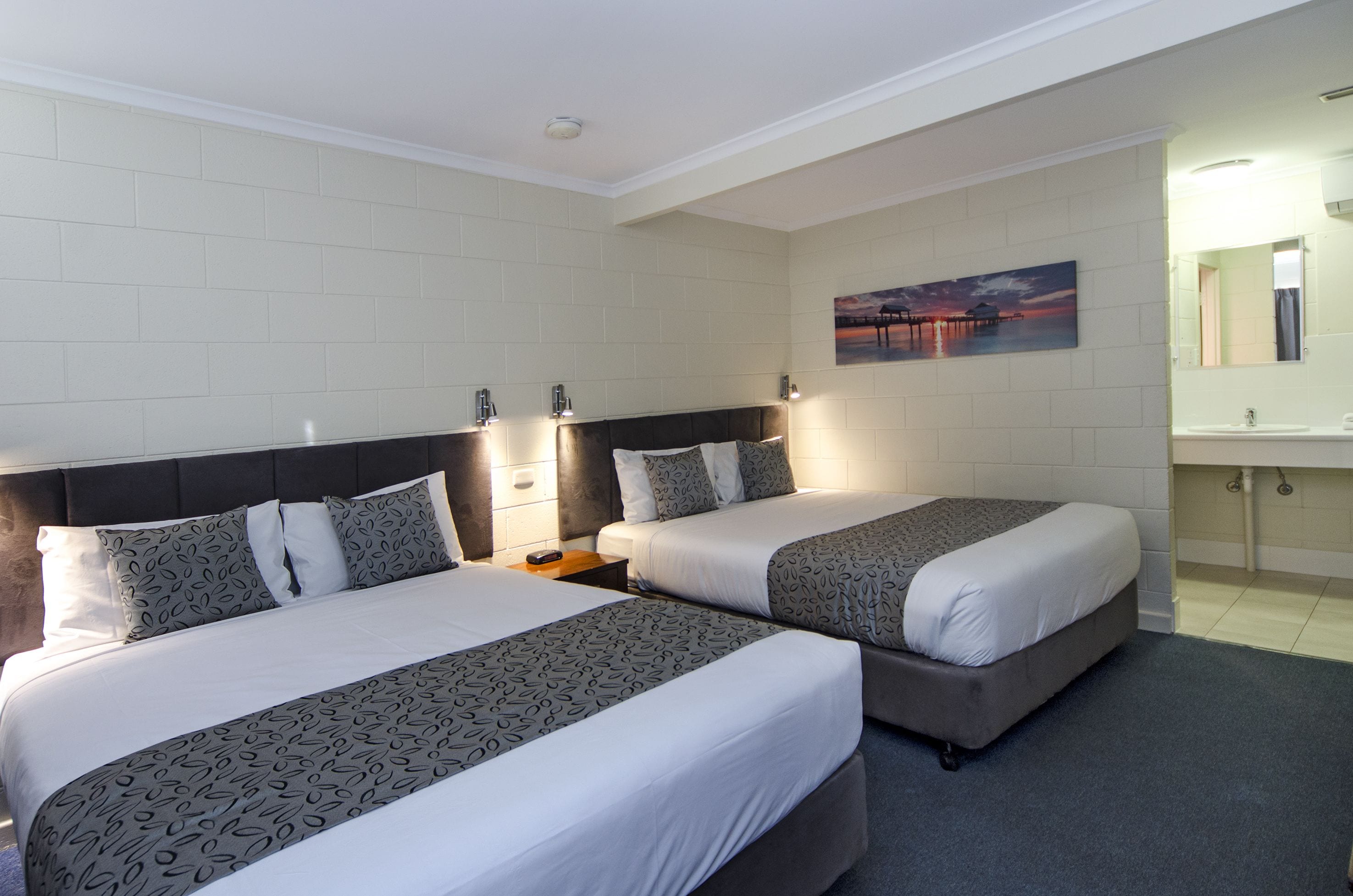 A hotel room with two beds, a nightstand, a painting, and a bathroom with a sink and mirror.