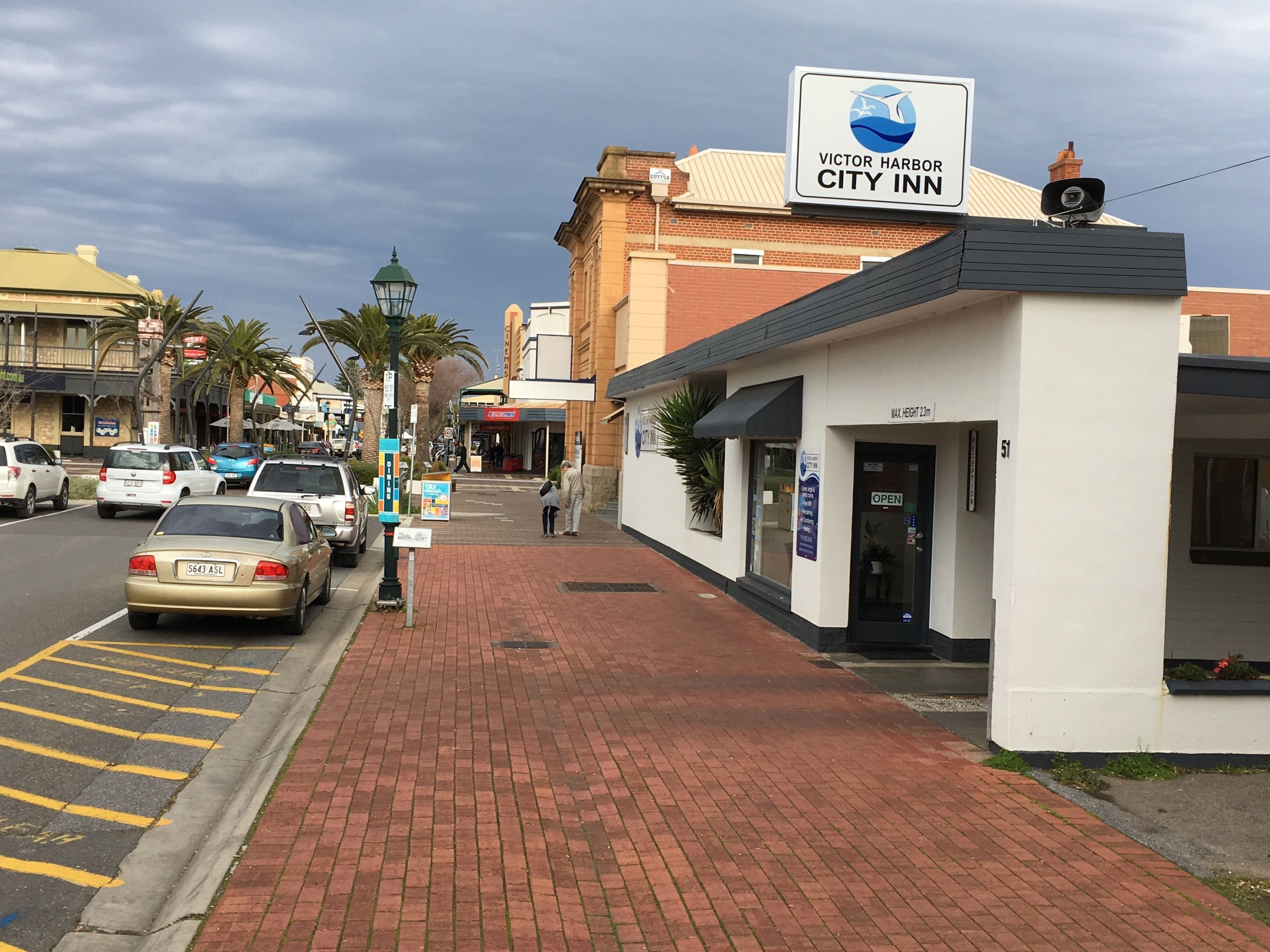 A city street with a Victor Harbor City Inn building, featuring a signboard, brick pavement, and parked cars.