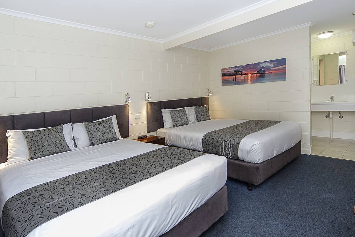 Quarto de hotel com duas camas, mesa de cabeceira, um quadro, banheiro com pia e espelho, e piso coberto por carpete.