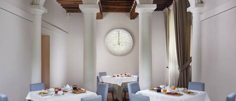 Un comedor con mesas blancas y sillas azules, un reloj redondo grande en la pared y vigas de madera en el techo.