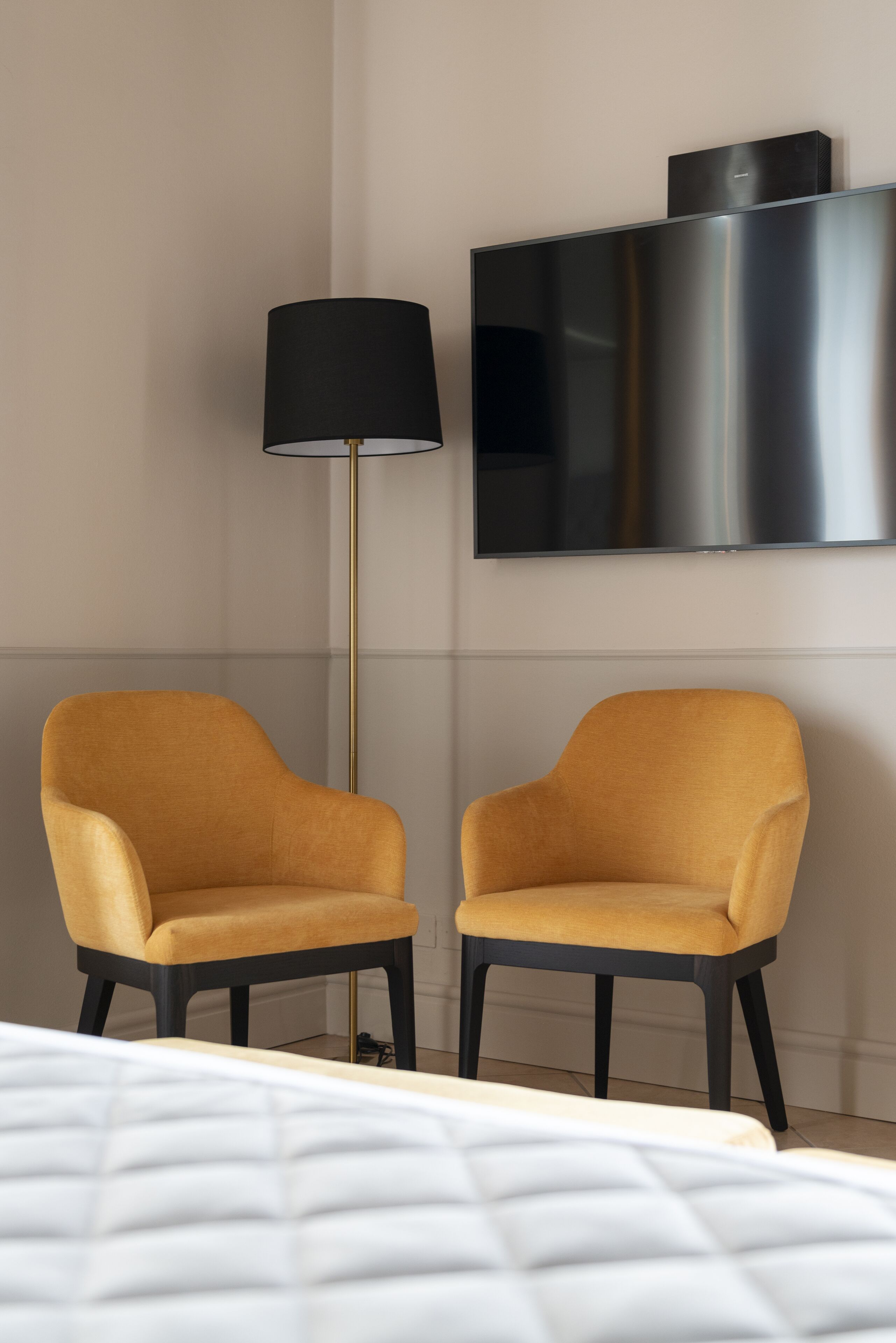 A modern hotel room with a bed, two mustard-colored armchairs, a floor lamp, and a flat-screen TV mounted on the wall.