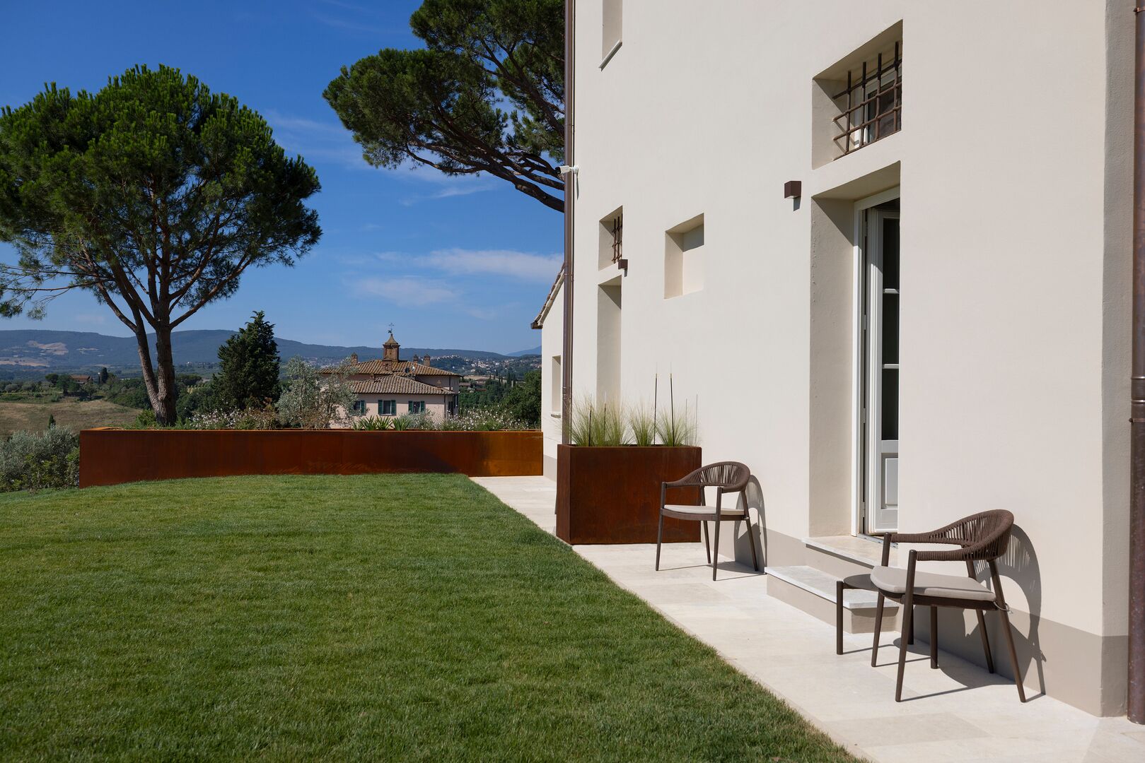 Una casa moderna con césped cuidado, dos sillas de exterior y vistas a edificios lejanos.