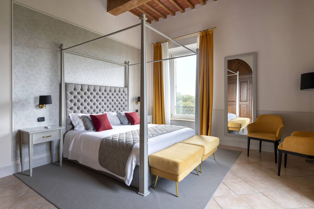 A four-poster bed with a tufted headboard, a bedside table with a lamp, a bench, and two chairs.