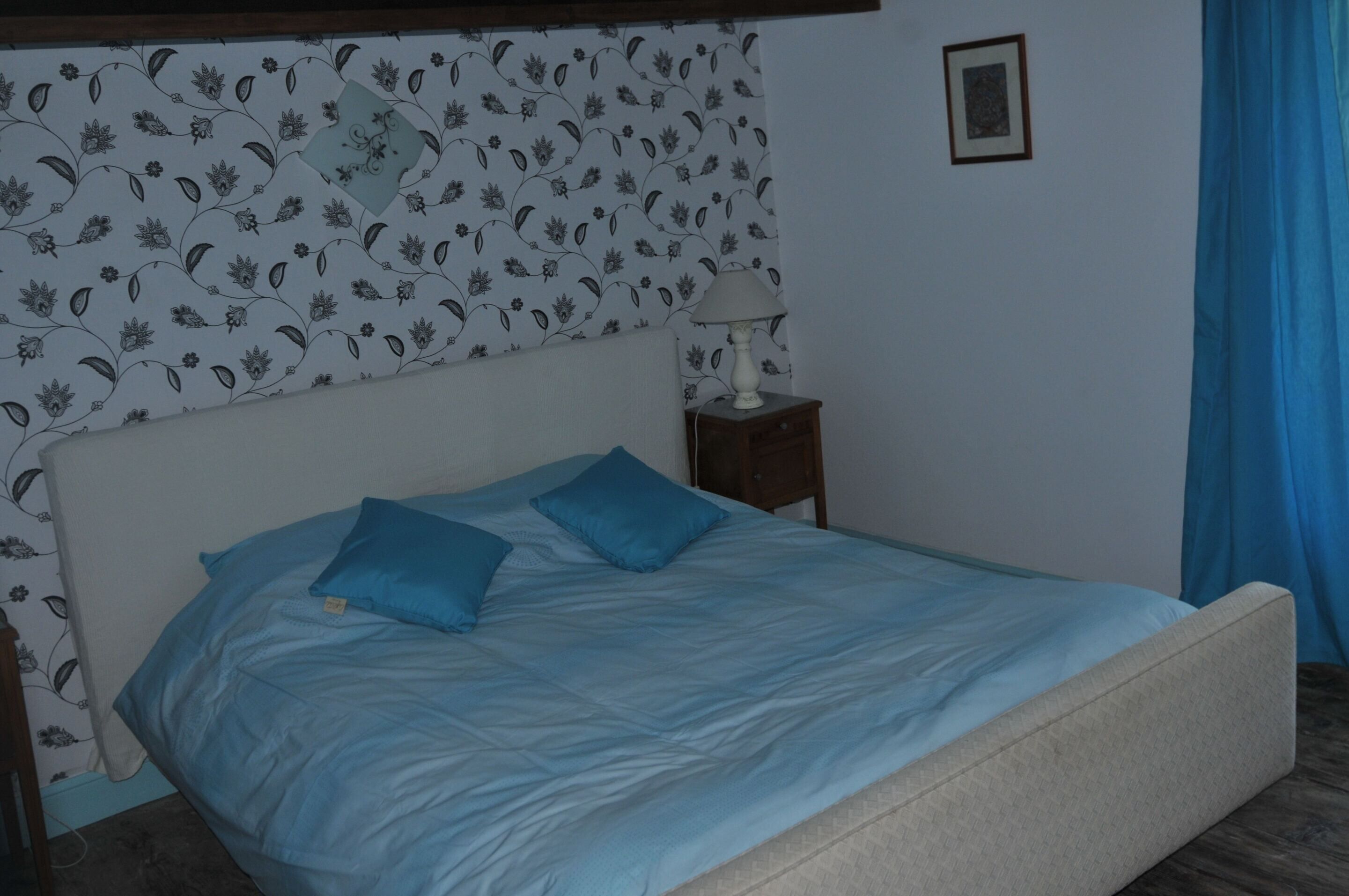 A bedroom with a bed, two pillows, a nightstand with a lamp, floral wallpaper, and a framed picture on the wall.