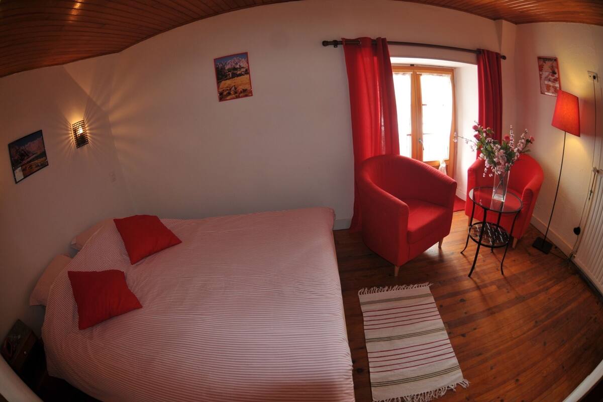 Una habitación con una cama, un sillón rojo, una mesita con un florero y una lámpara.