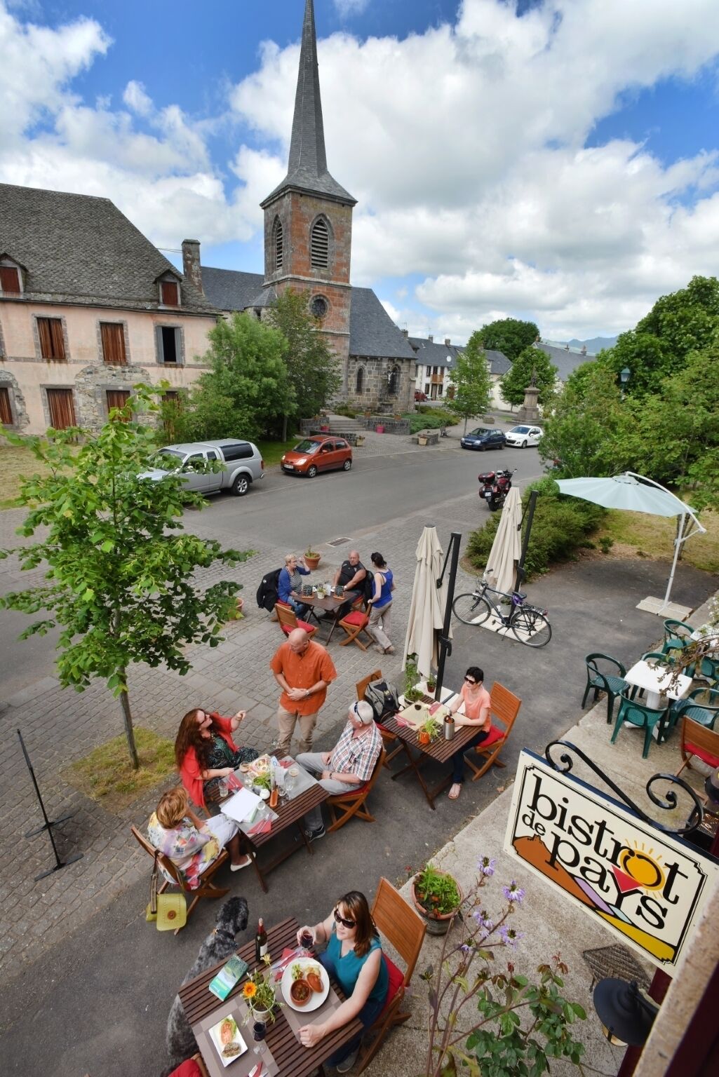 Un espace extérieur aménagé pour les repas, avec des tables et des chaises, une église en arrière-plan et une enseigne indiquant « Bishop de Pays ».