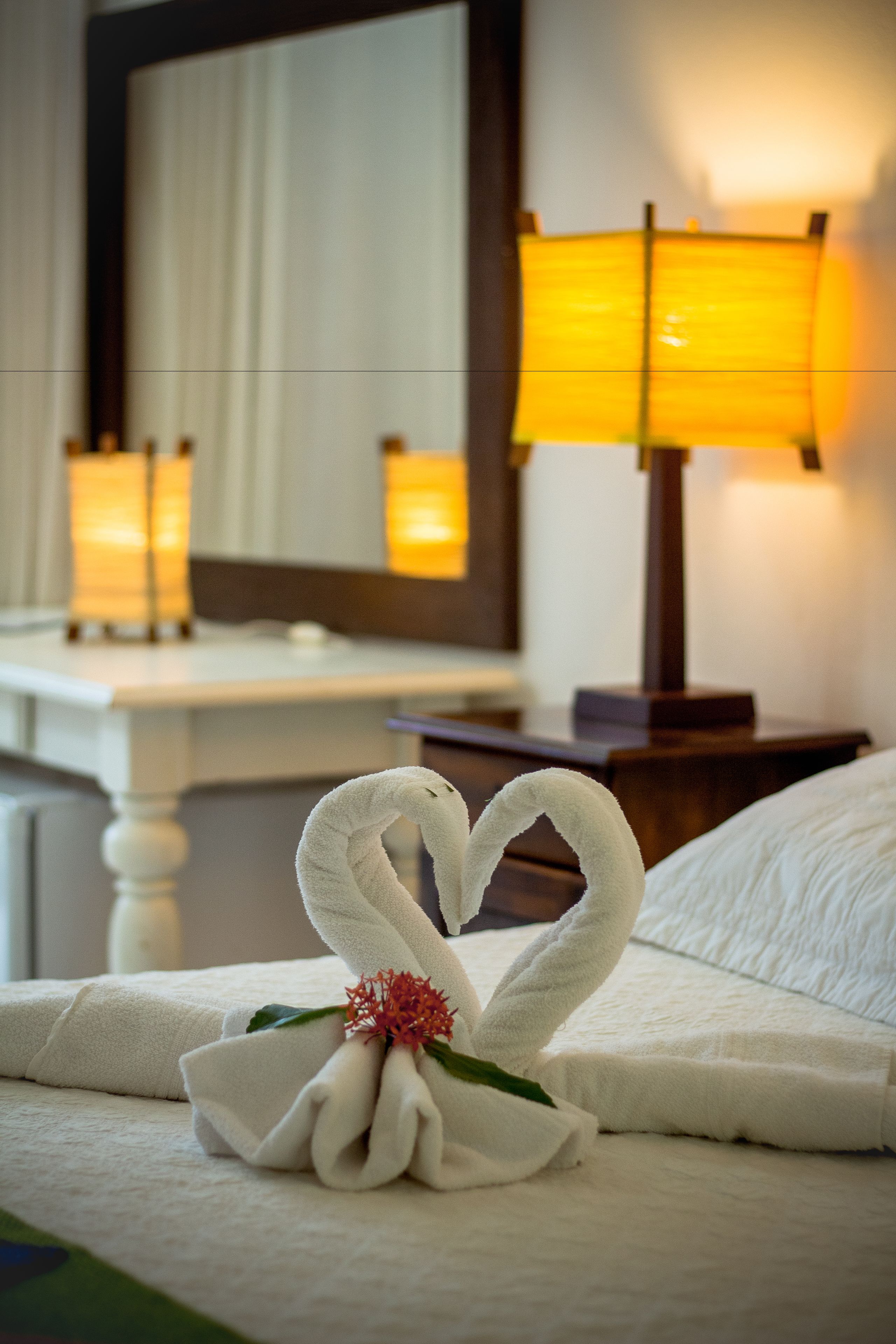 Una cama bien hecha con una decoración de cisne de toallas, una lámpara de noche y un espejo que refleja la decoración de la habitación.