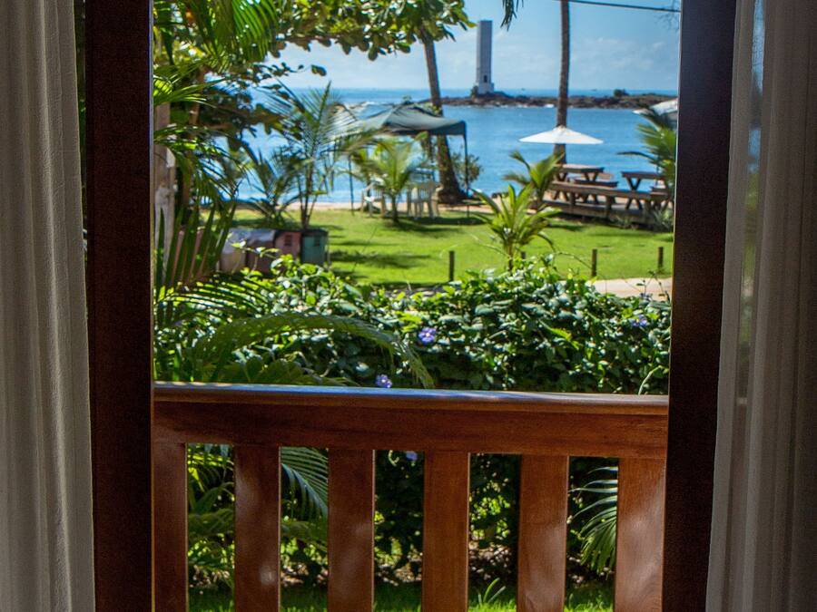 Une vue depuis un balcon doté d’une balustrade en bois, d’un hamac en tissu et d’un jardin luxuriant, verdoyant, avec des palmiers et une étendue d’eau en arrière-plan.
