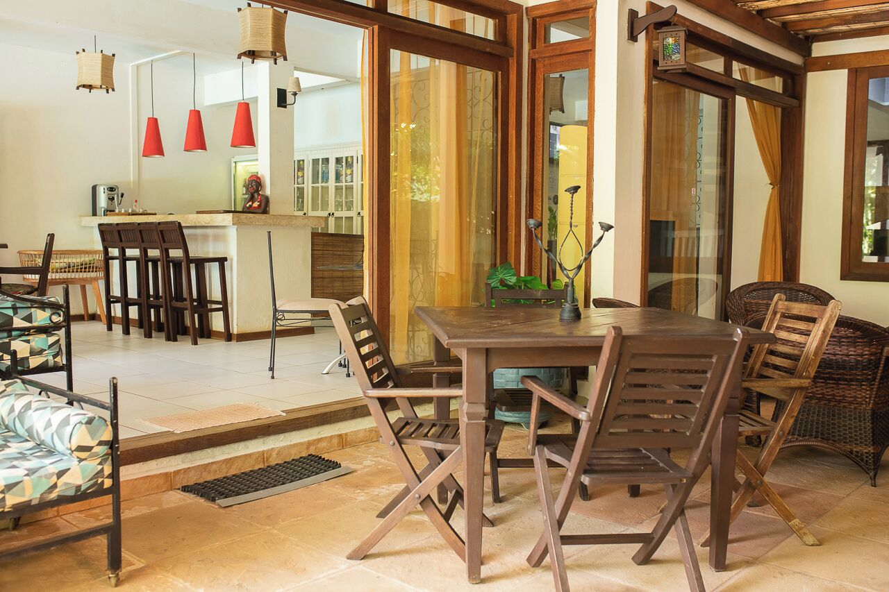 A dining area with a wooden table and chairs, a kitchen in the background, and a large window with curtains.