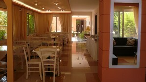 A modern dining area with a glass table and chairs, a kitchen with a fan, and a sofa by a window. - Avra Hotel (Lefkada)