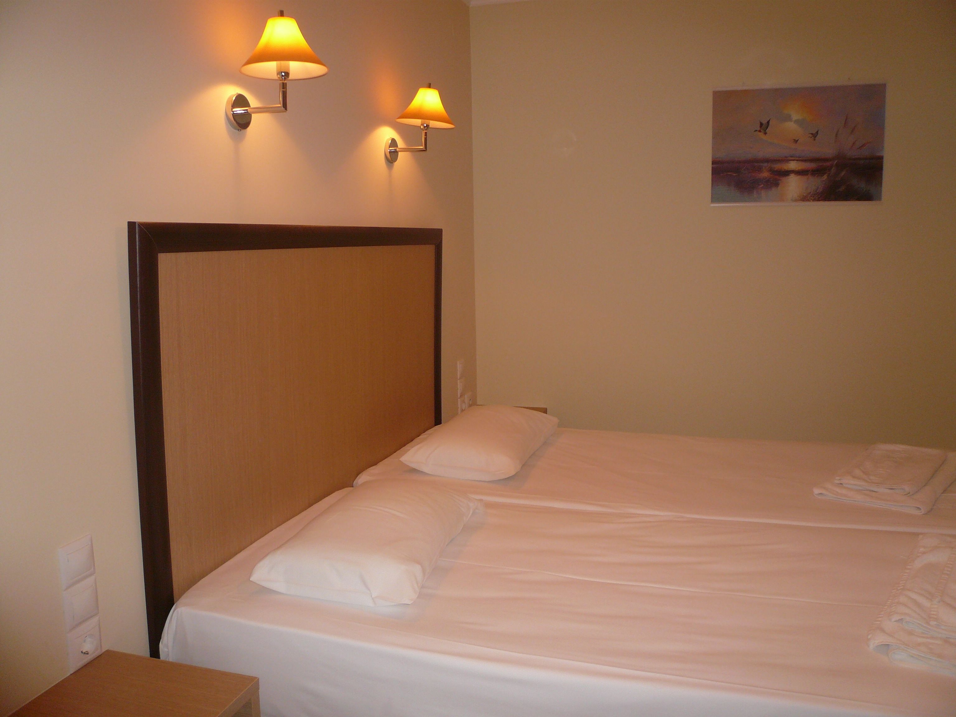 A hotel room with a wooden headboard, two bedside lamps, a painting of a sunset, and a neatly made bed with white linens.