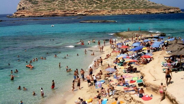 A crowded beach with people swimming and sunbathing, a rocky hill in the background, and clear blue water. - Apartamentos Tramuntana (Sant Antoni de Portmany)