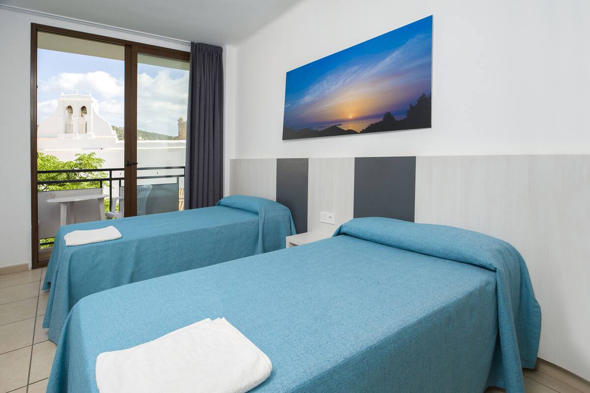 A hotel room with two beds, a balcony, a painting of a sunset, and a view of a white building.