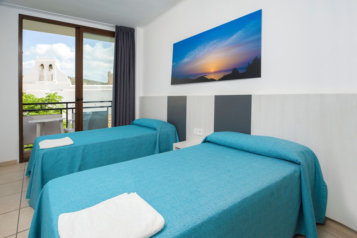 A hotel room with two beds, a balcony, a painting of a sunset, and a view of a white building.