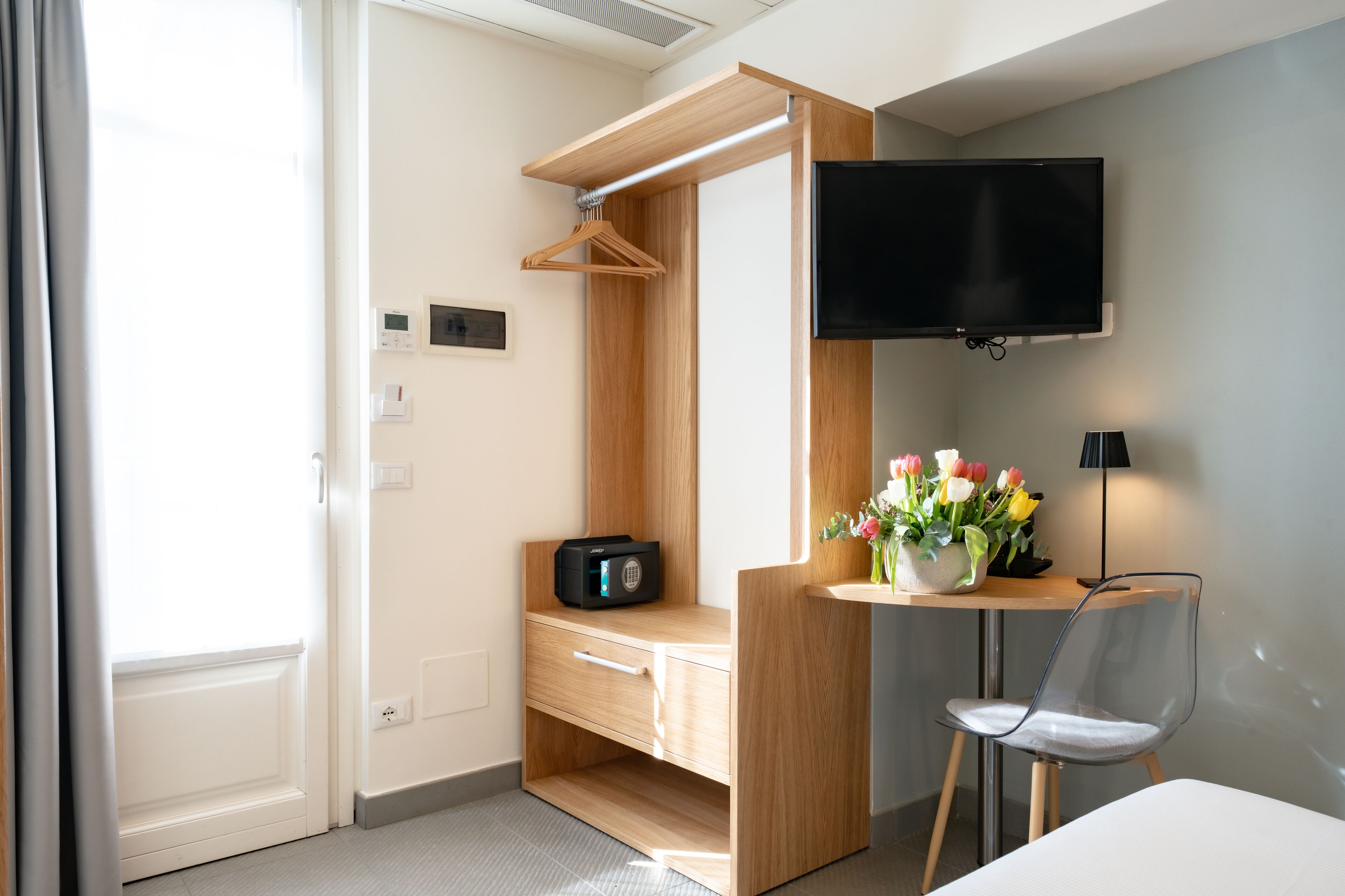 A compact hotel room with a wooden desk, a TV mounted on the wall, a chair, and a small table with flowers.