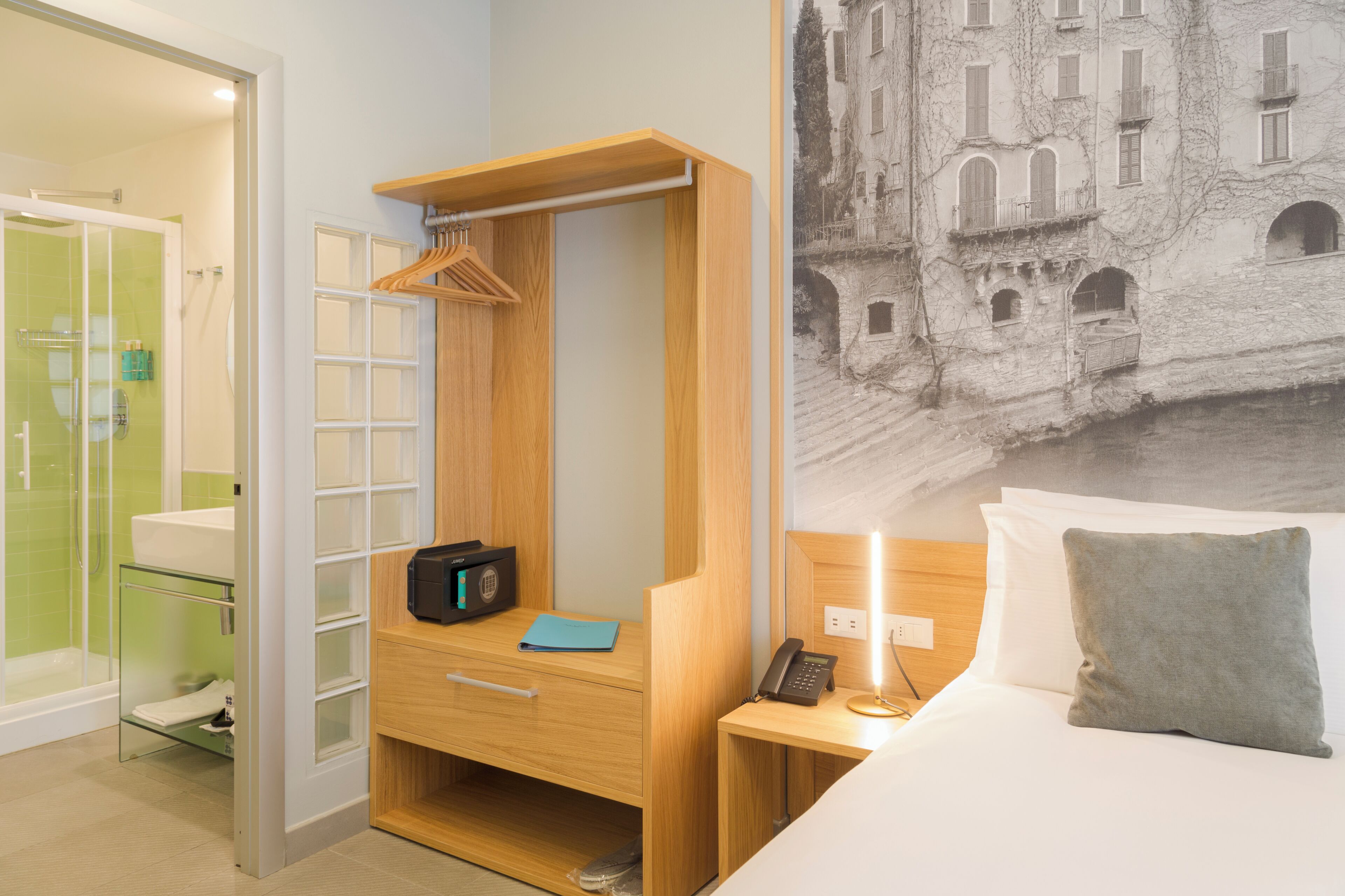 A hotel room with a wooden wardrobe, a bedside table with a lamp, a bed with a pillow, and a wall-mounted telephone.