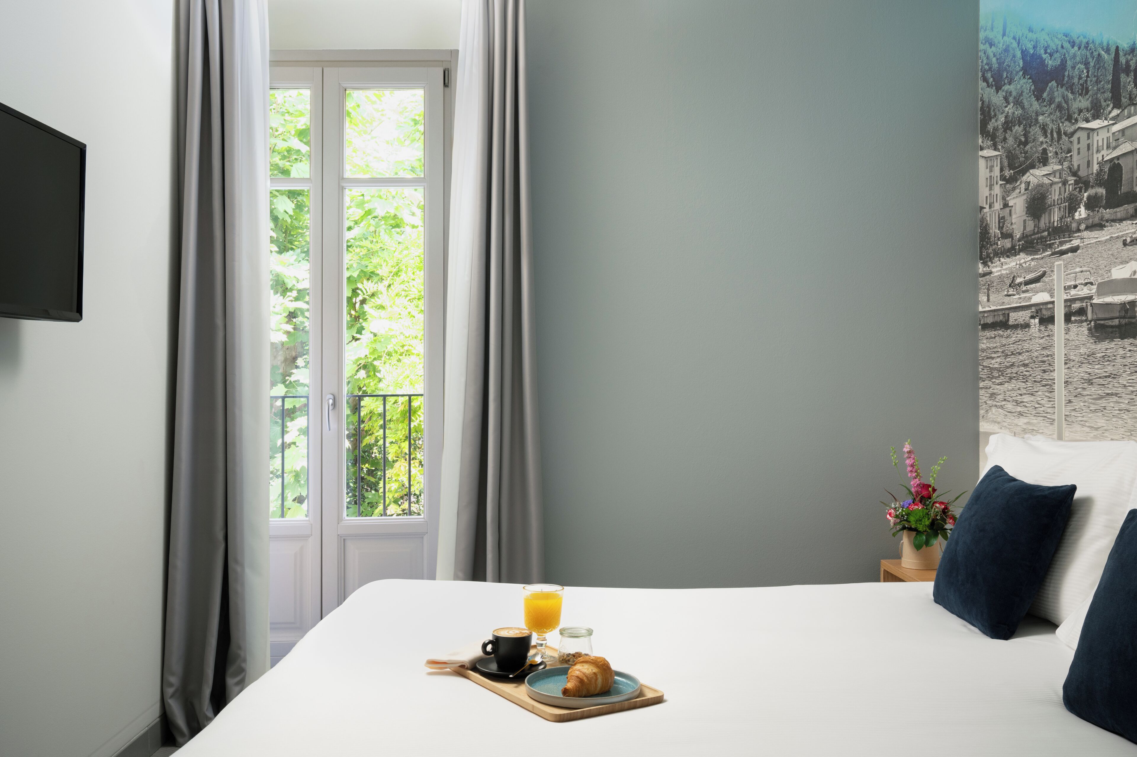 Habitación de hotel con cama, bandeja de desayuno con croissant, café y zumo de naranja, mesita de noche con un florero y vista a la ciudad a través de la ventana.