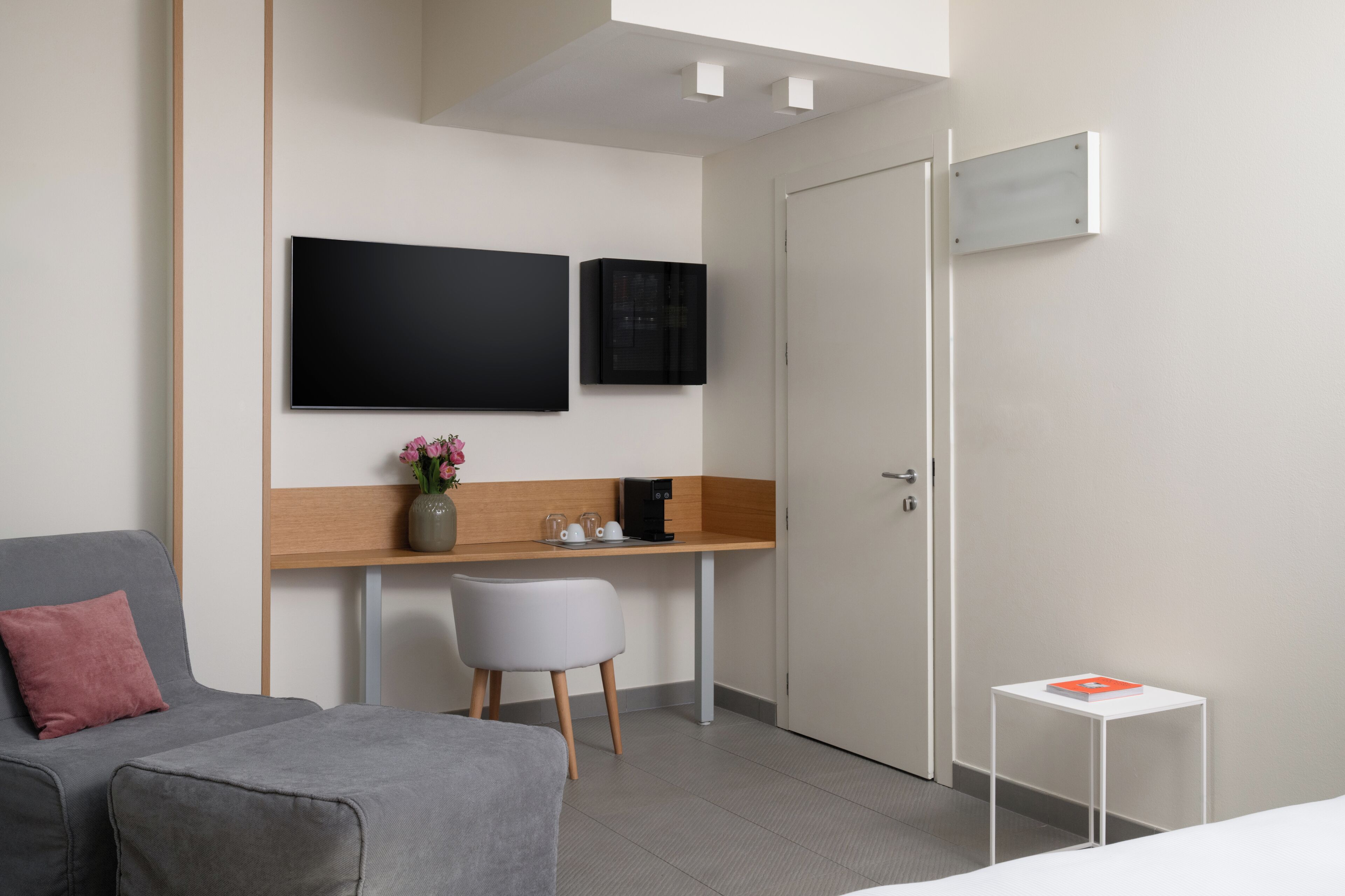 Moderni hotellihuone, jossa on harmaa sohva, pieni valkoinen pöytä, työpöytä tietokoneella ja maljakko kukkineen sekä seinälle asennettu litteänäyttöinen televisio.