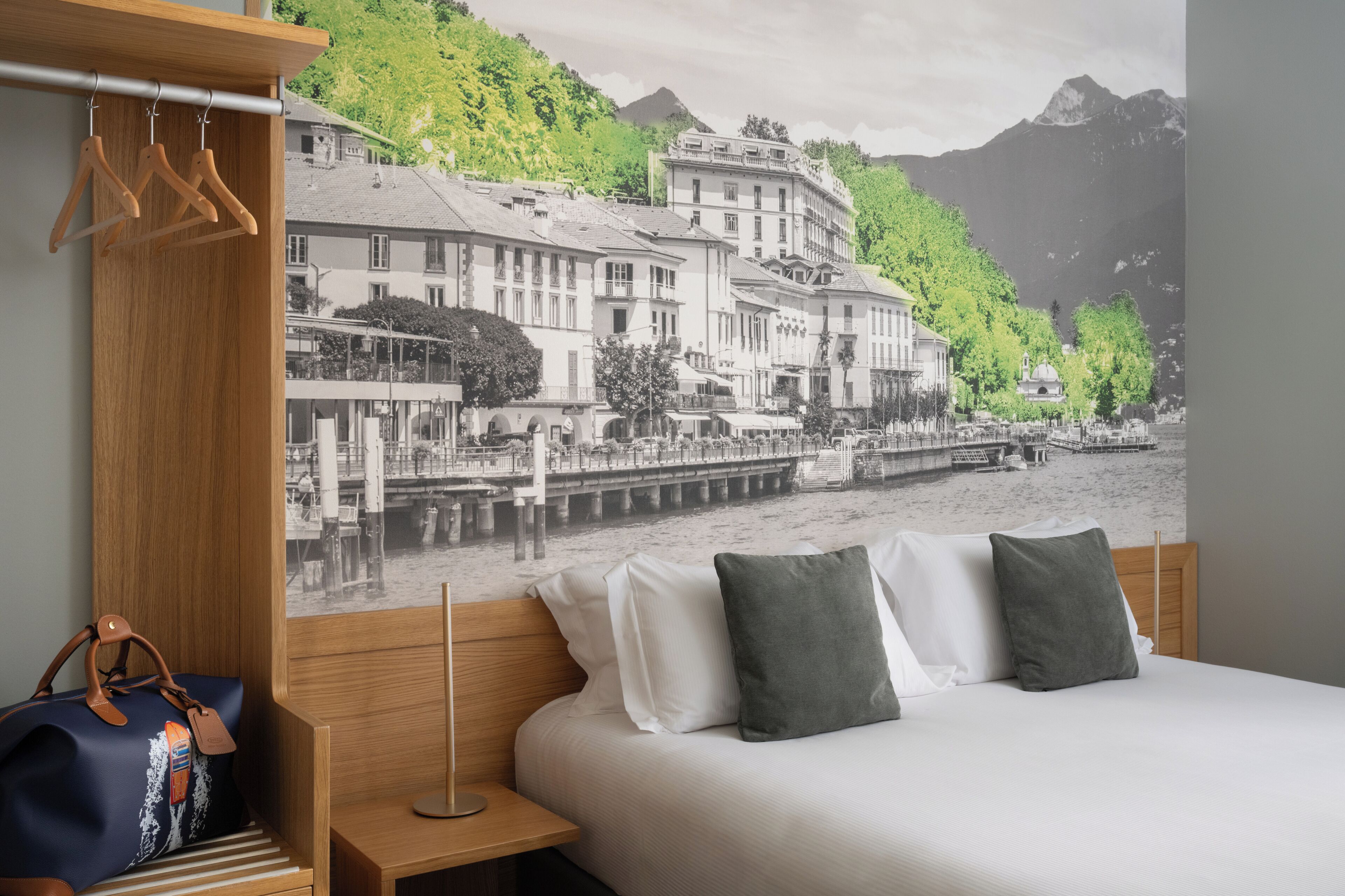 A hotel room with a bed, a wooden nightstand, a hanging coat rack, and a wall mural of a riverside town.