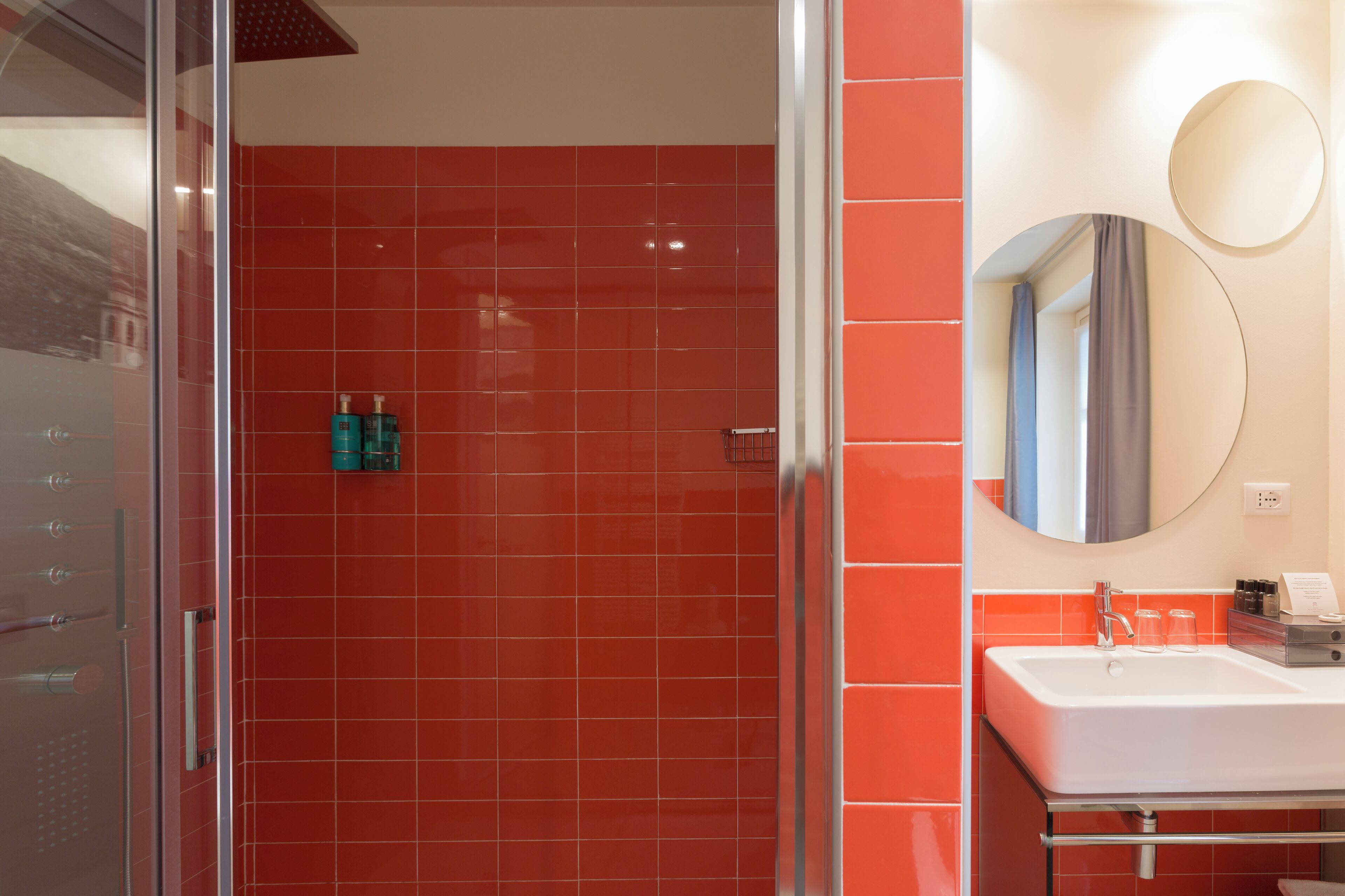 Un baño moderno con baldosas rojas, una cabina de ducha de vidrio, un lavamanos blanco y un espejo redondo.