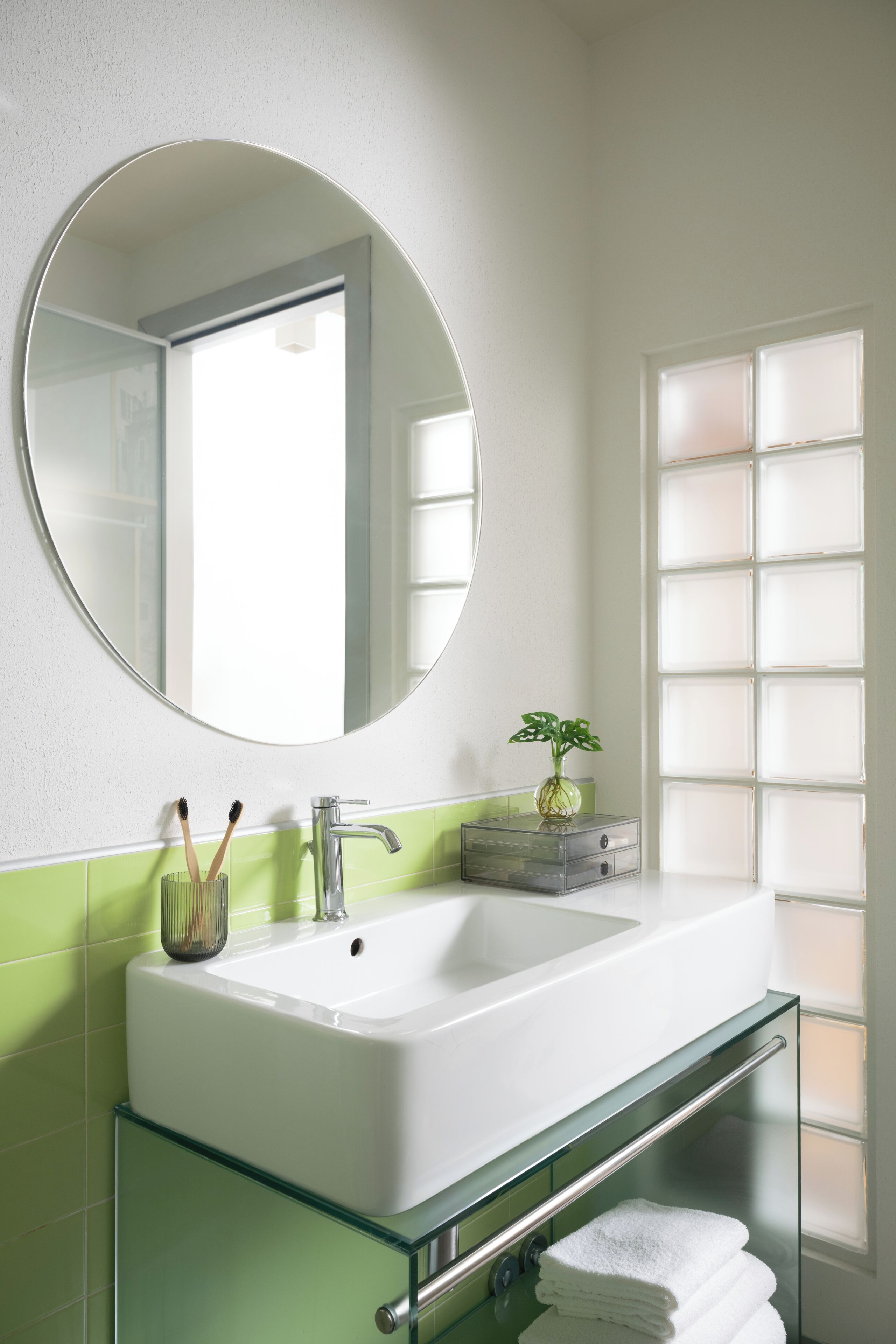 Un bagno con un lavandino bianco, piastrelle verdi, uno specchio rotondo e una finestra con vetro satinato.