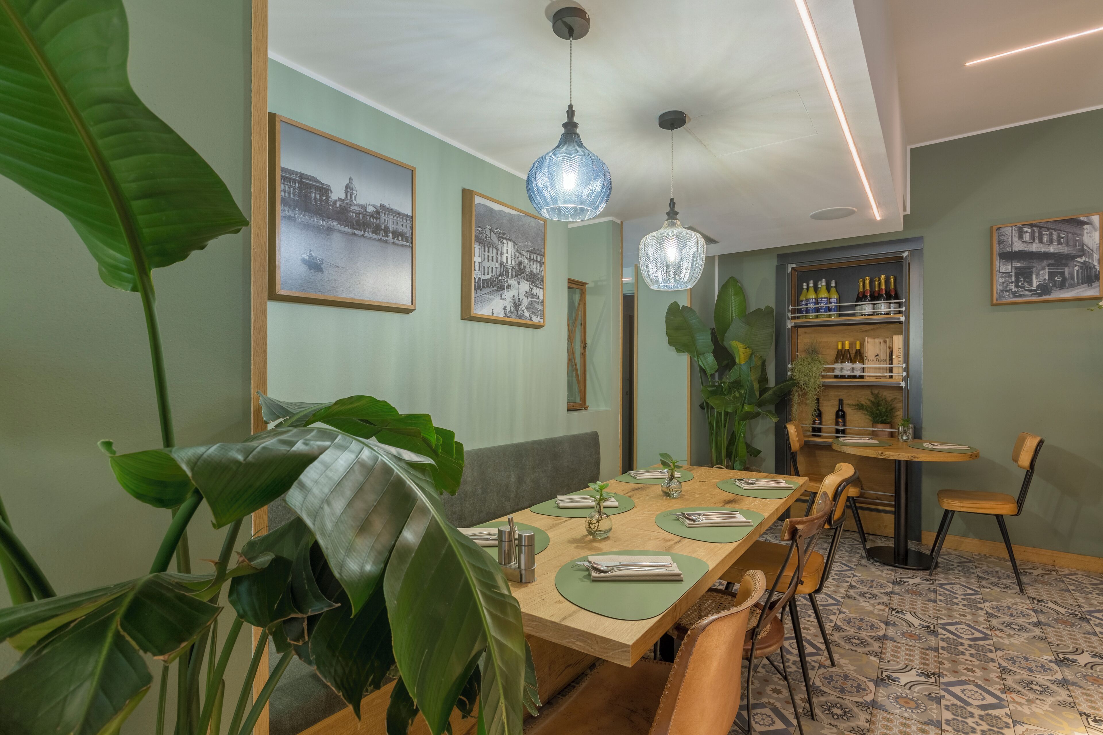 A dining area with a long wooden table, green chairs, and a bench. There are framed pictures on the walls, a hanging light fixture, and a shelf with bottles.