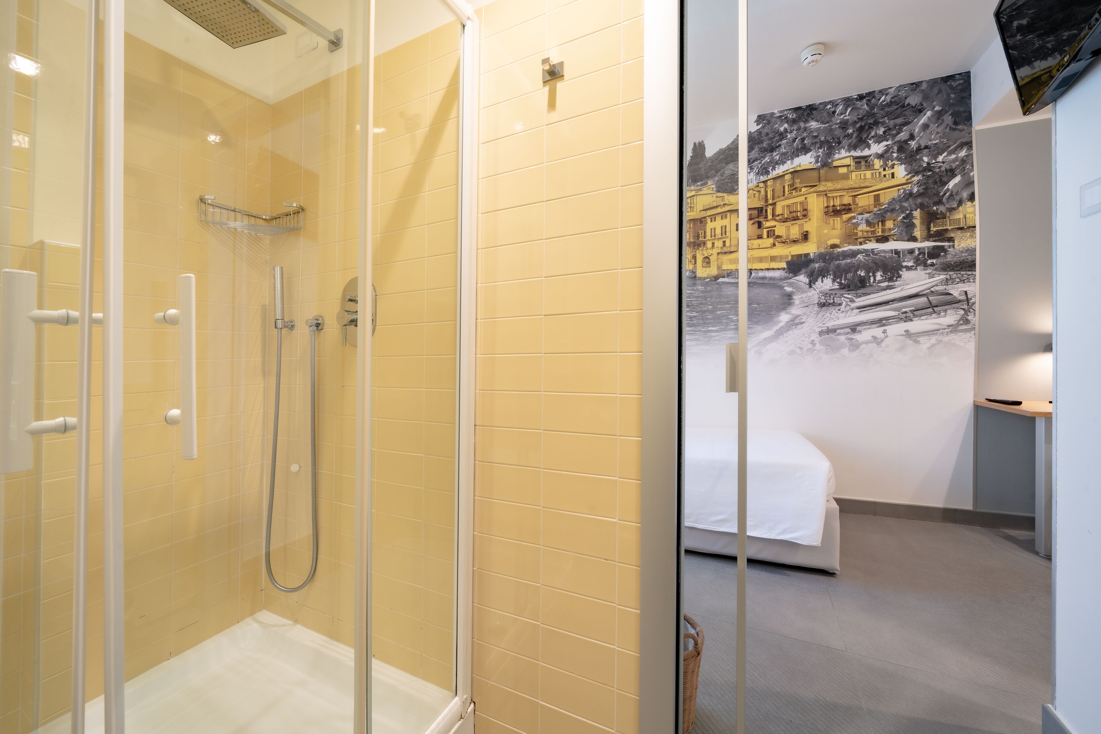 Une chambre d’hôtel dotée d’une douche en verre, d’un lit avec une literie blanche et d’une fresque représentant un paysage côtier.