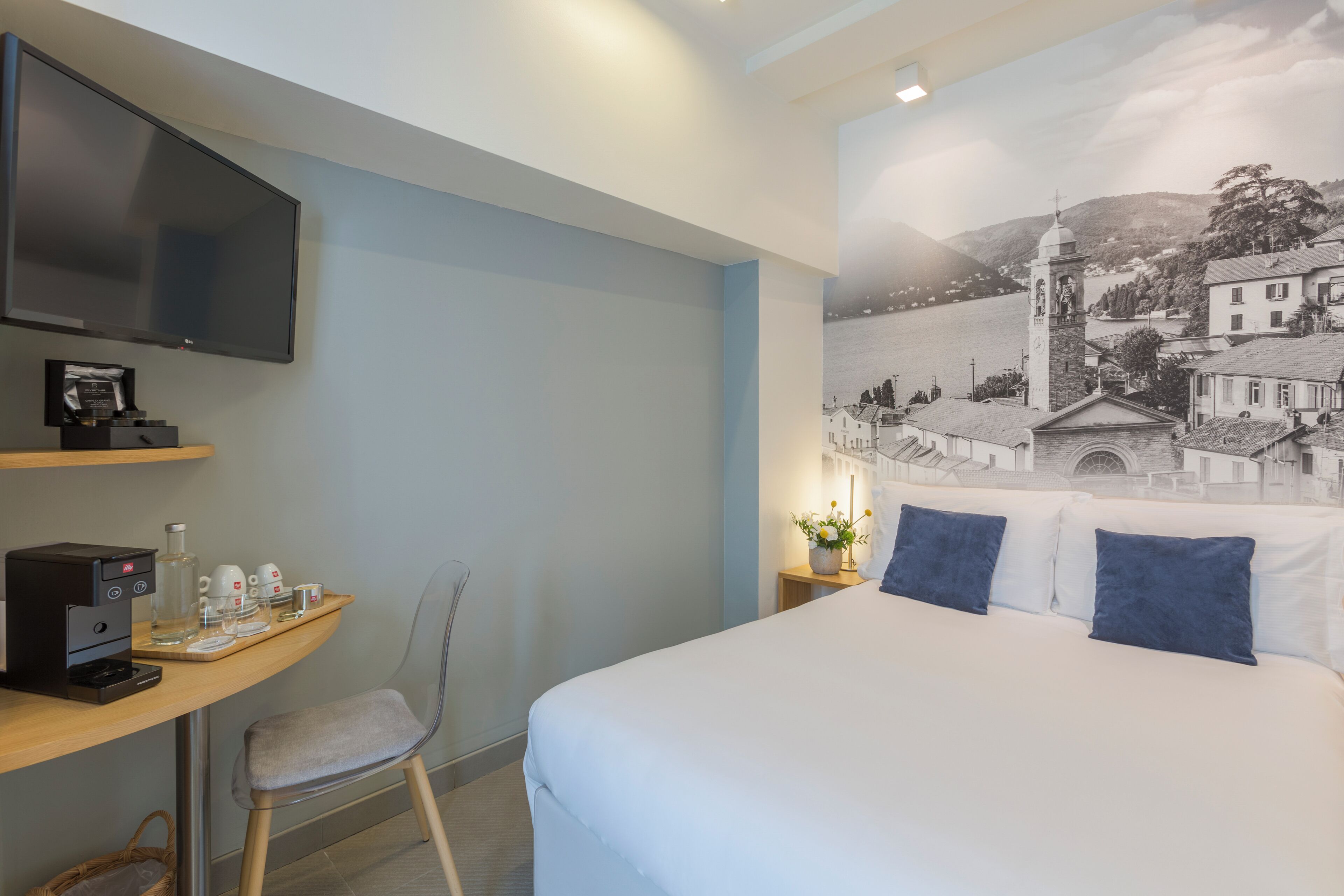 A hotel room with a bed, a desk with a coffee machine, a chair, and a wall-mounted TV.