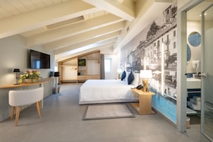 A modern hotel room with a large bed, a desk with a flat-screen TV, a seating area with a chair, and a bathroom with a towel rack.