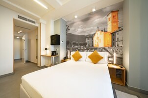 A hotel room with a large bed, a nightstand with a lamp, a wall-mounted TV, and a mural of a town scene.