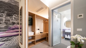 Ein modernes Hotelzimmer mit einem Holzschrank, einem Bett mit violettem Baldachin, einem kleinen Schreibtisch mit Lampe sowie einem Badezimmer mit Waschbecken und Spiegel.