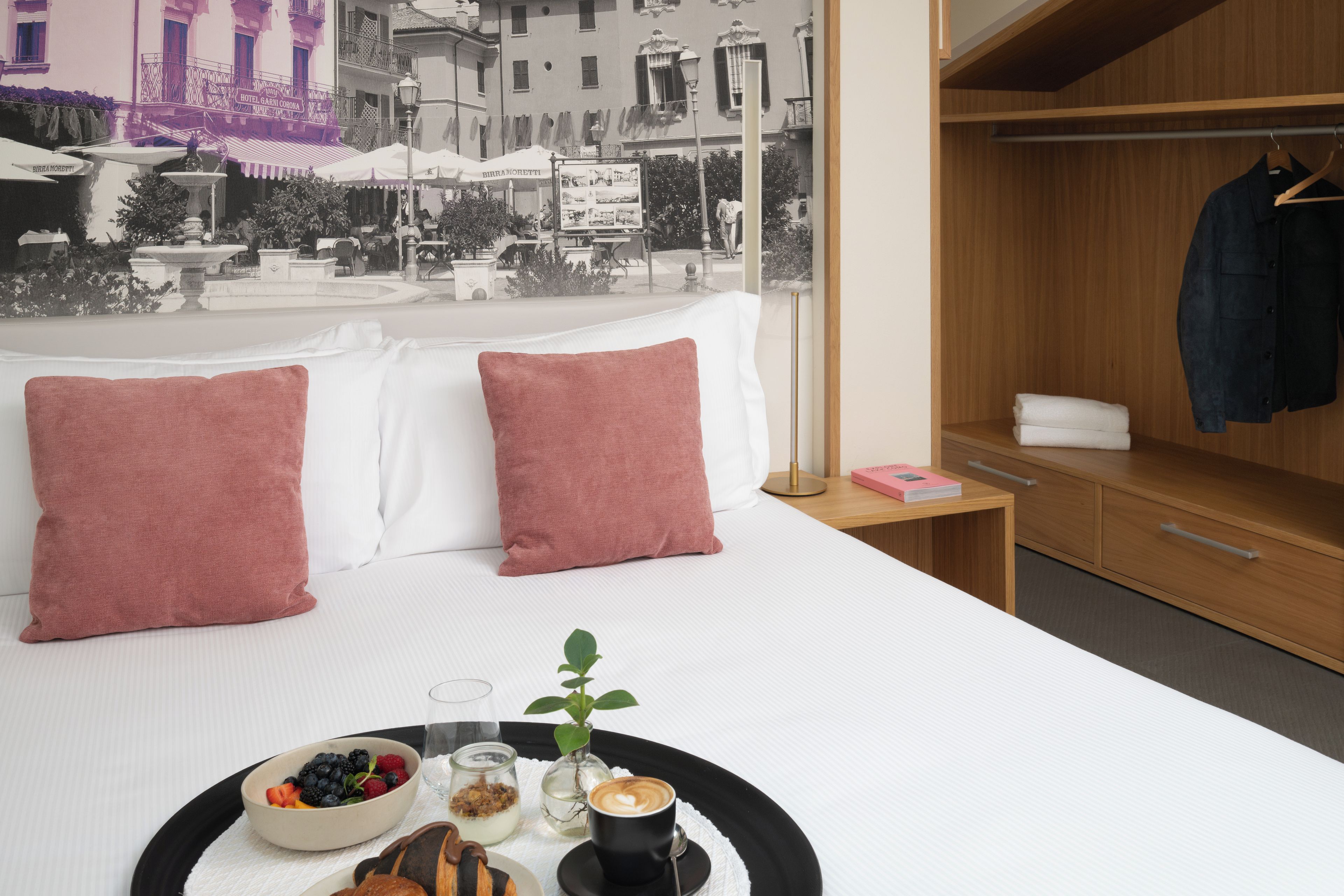 A hotel room with a bed, a bedside table with a tray of breakfast, a window with a view of a cityscape, and a wooden wardrobe.
