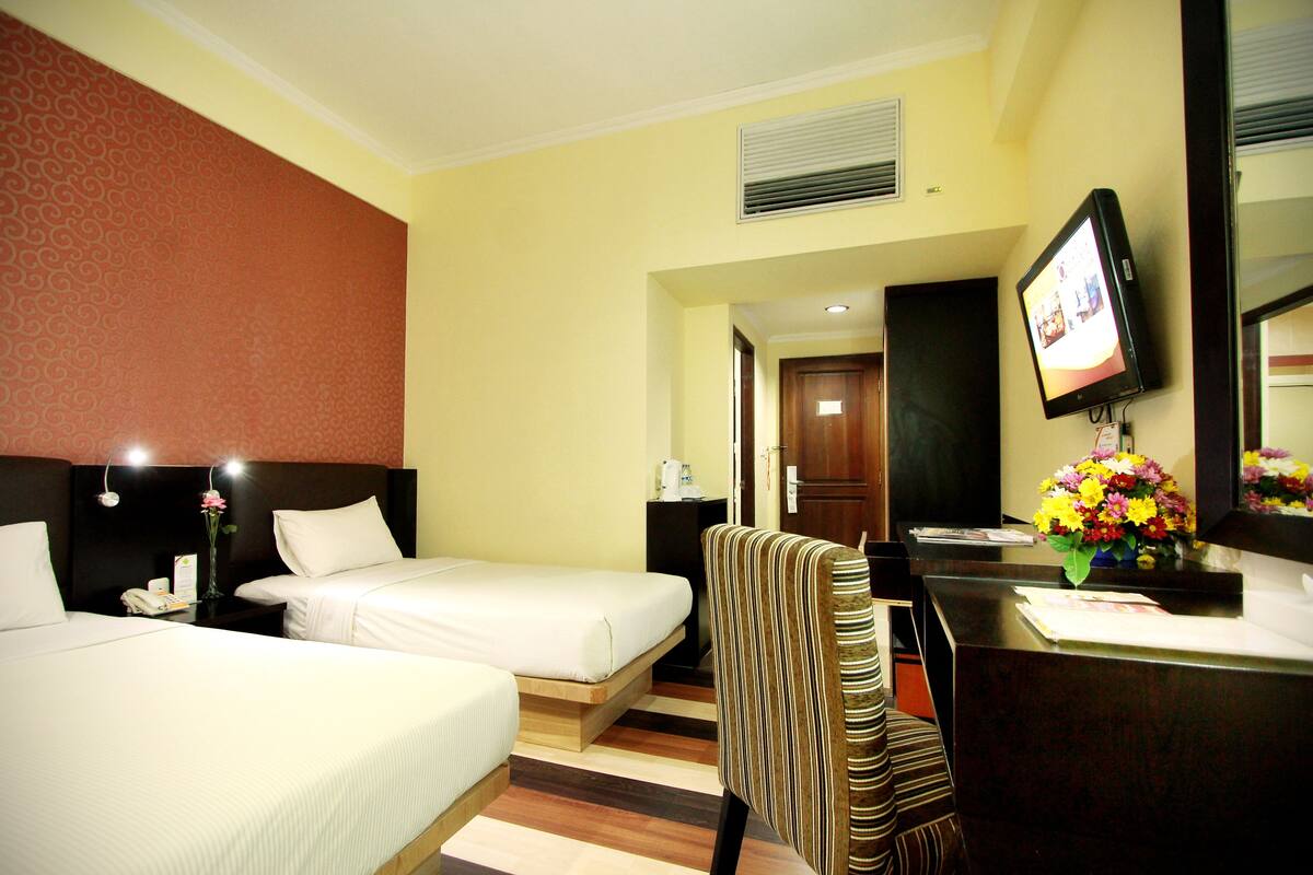 A hotel room with two beds, a desk with a computer, a chair, a TV, and a flower vase.