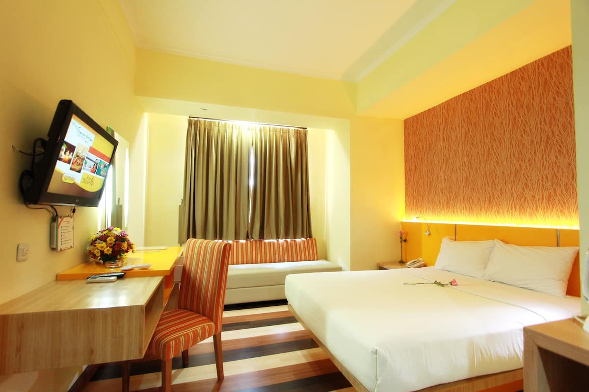 A hotel room with a bed, a desk with a TV, a chair, a sofa, and a window with curtains.