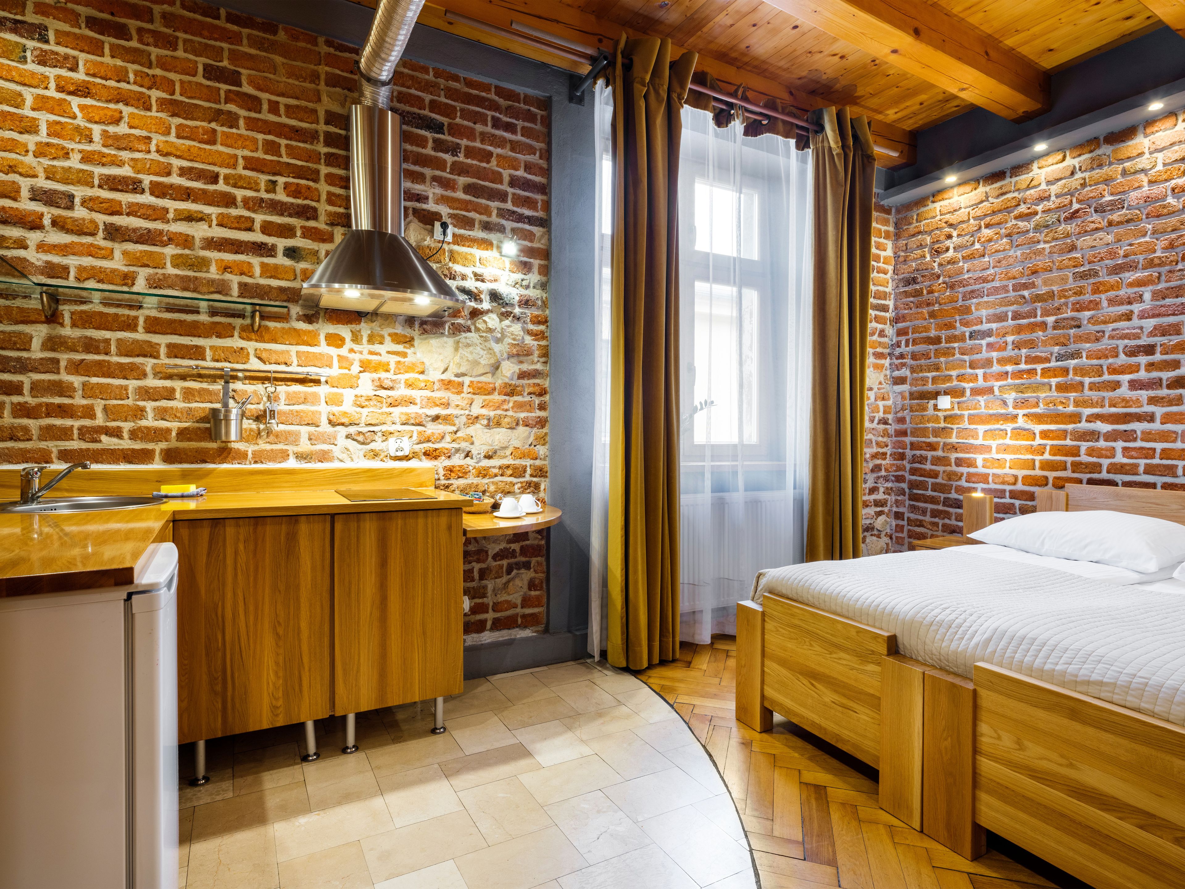 Een knus kamertje met een bakstenen muur, een houten bed, een klein houten bureau met een wastafel en een raam met gordijnen.