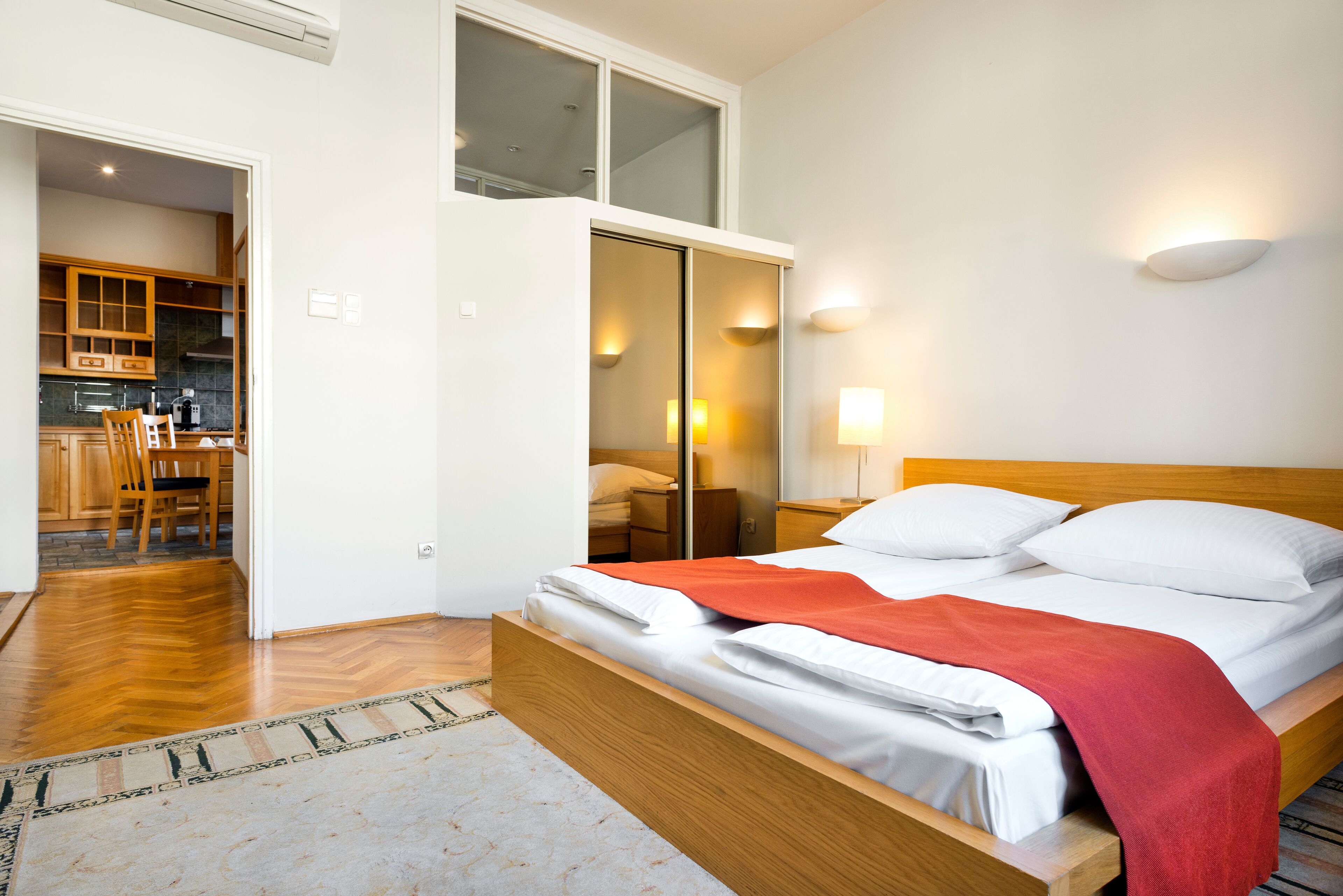 A bedroom with a wooden bed, a red throw blanket, and a view of a kitchen and dining area through an open door.