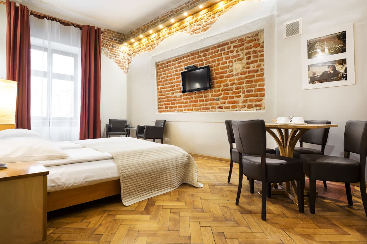 A hotel room with a bed, a desk, a table with chairs, a TV mounted on a brick wall, and a window with curtains.