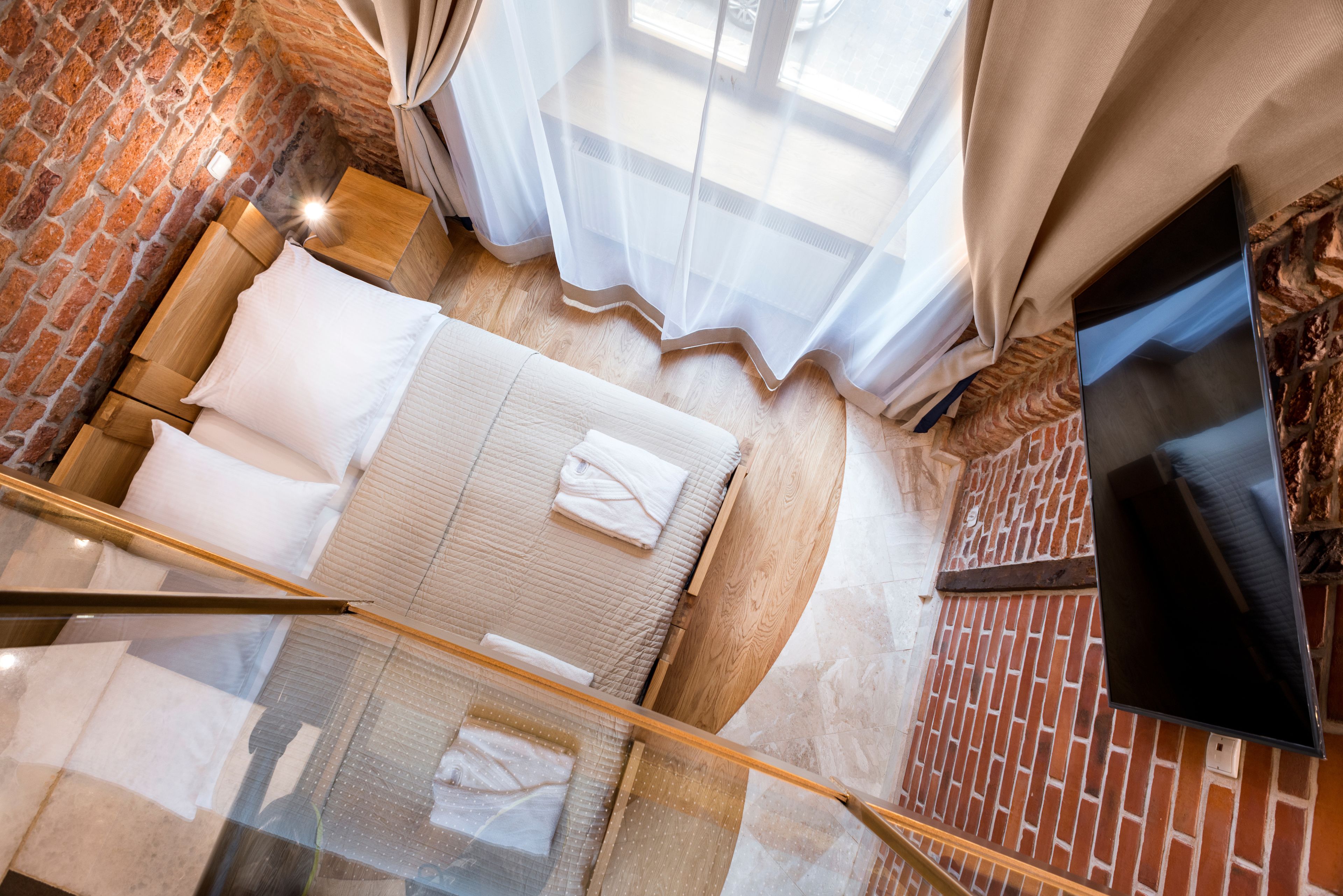 A hotel room with a brick wall, a bed with white linens, a TV on the wall, and a window with sheer curtains.