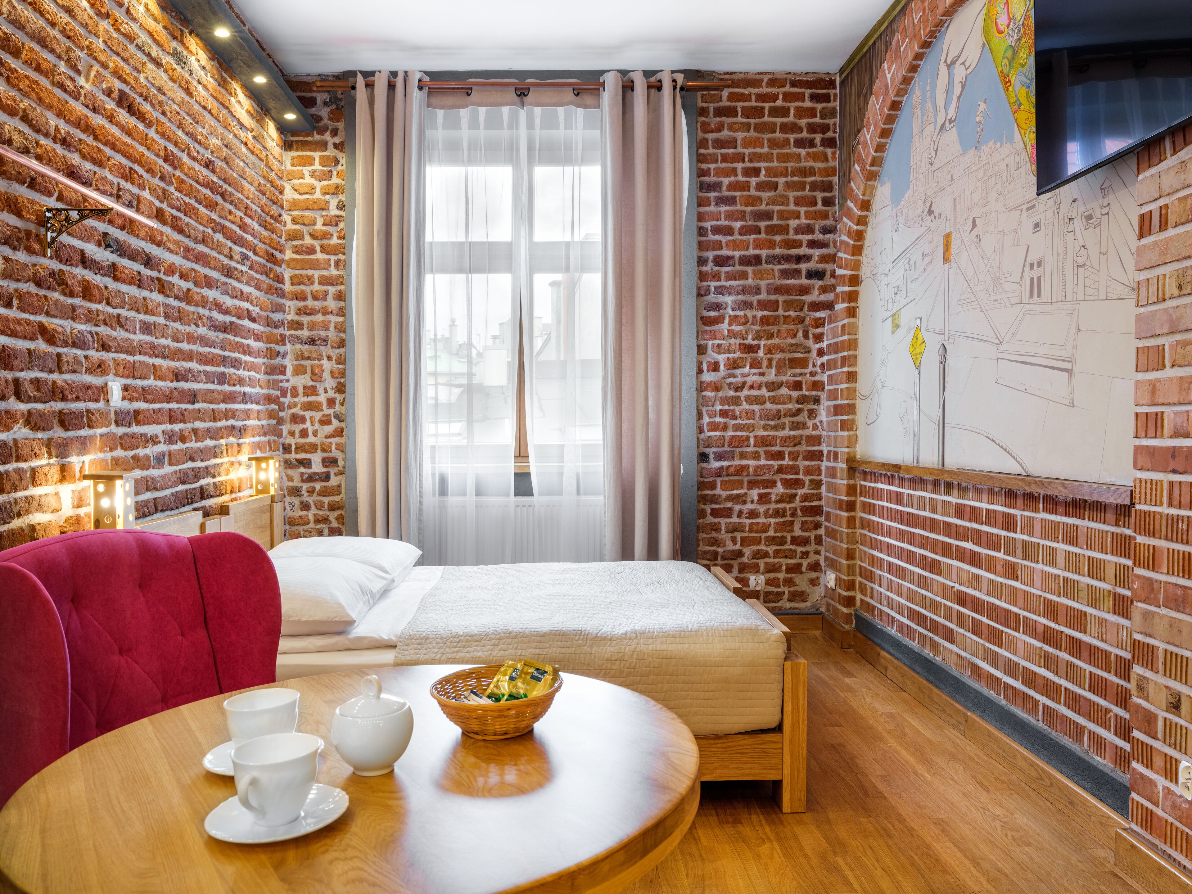 A room with a brick wall, a bed with white bedding, a red chair, a wooden table with cups and a basket, and a window with sheer curtains.