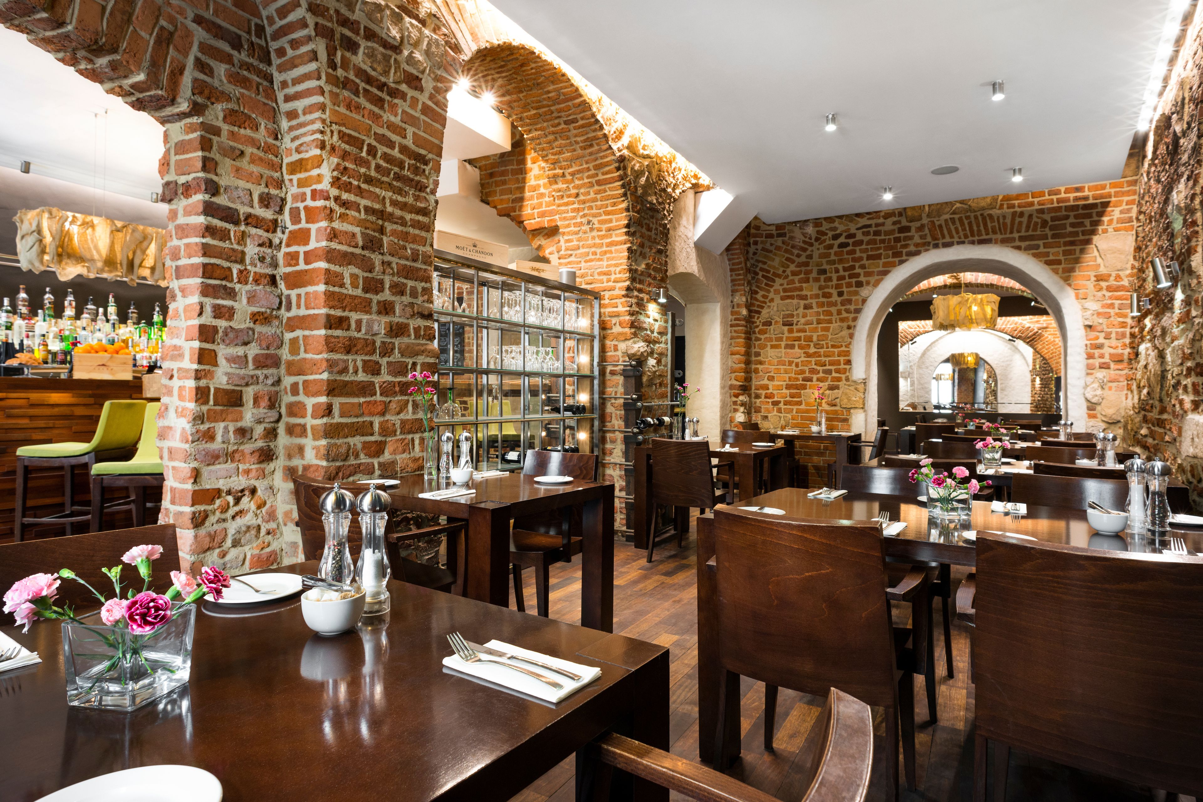 A restaurant with brick walls, wooden tables, and chairs. There are bottles and glasses on the shelves.