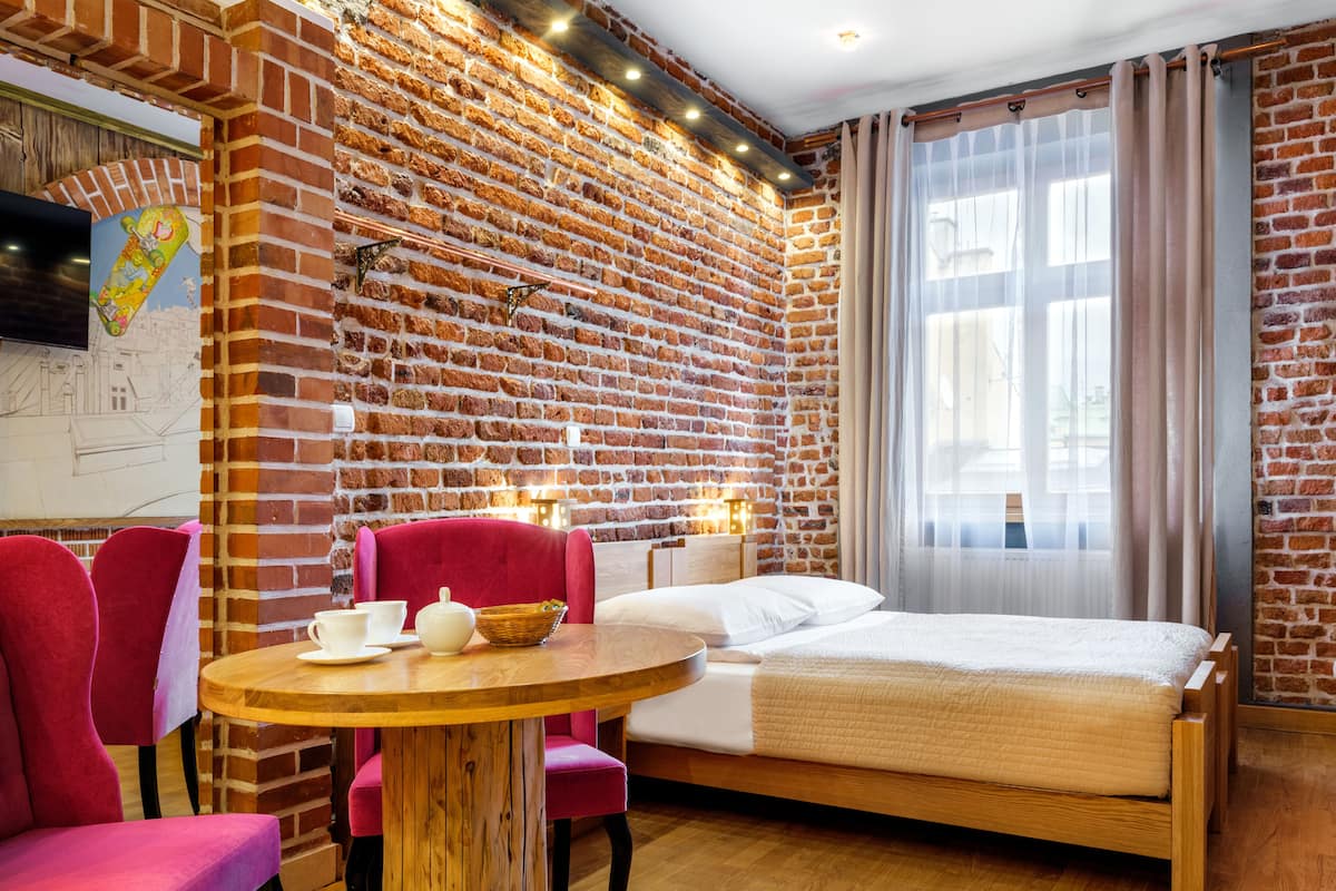A room with a brick wall, a bed, a round wooden table with chairs, and a TV on the wall.