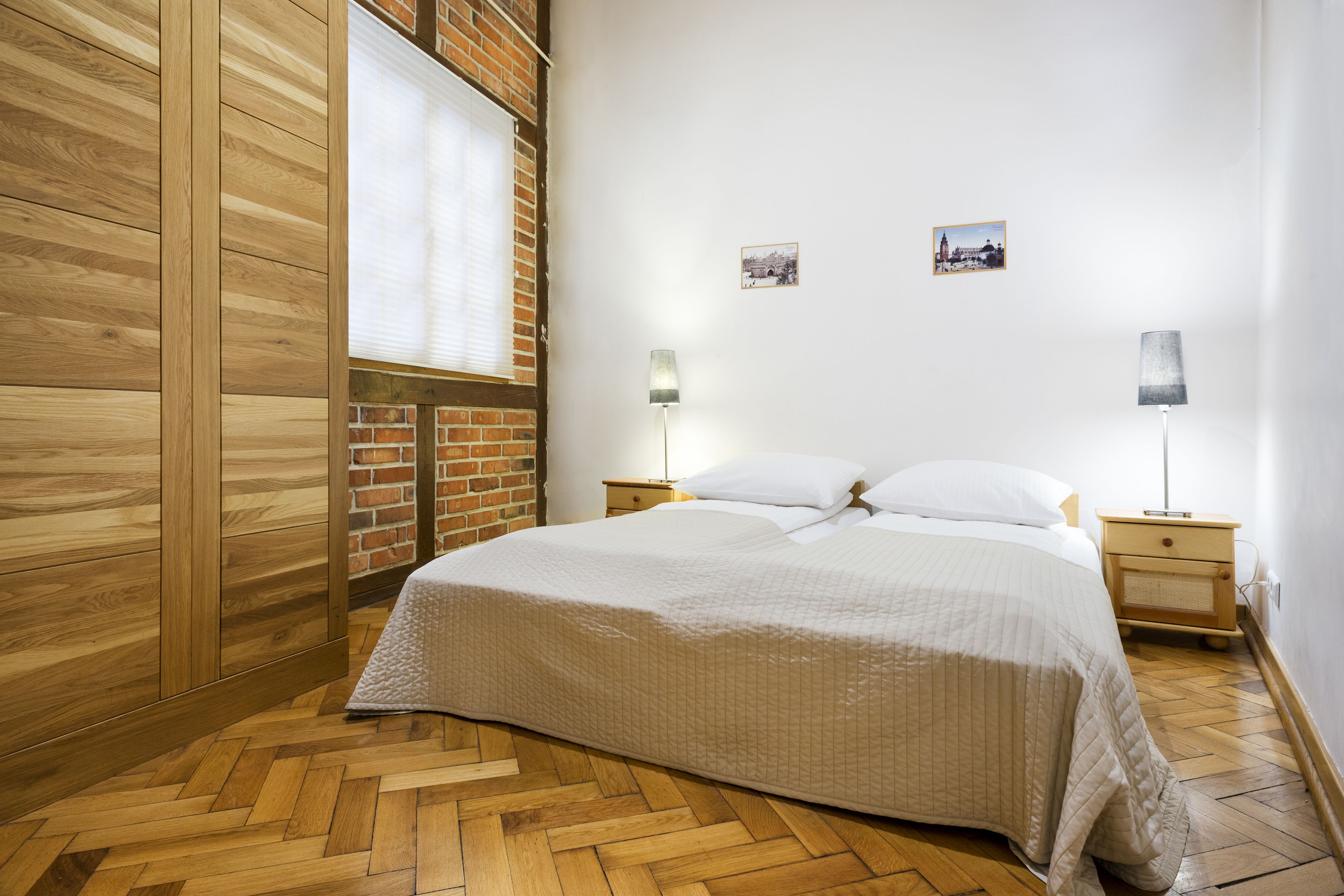 Quarto com chão de madeira, cama com roupa de cama branca, duas mesas de cabeceira com abajures e uma parede de tijolos.