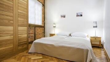Quarto com chão de madeira, cama com roupa de cama branca, duas mesas de cabeceira com abajures e uma parede de tijolos.