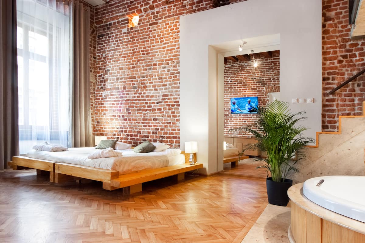 A modern bedroom with a brick wall, wooden flooring, a bed with pillows, a bedside table, a TV, a plant, and a bathtub.