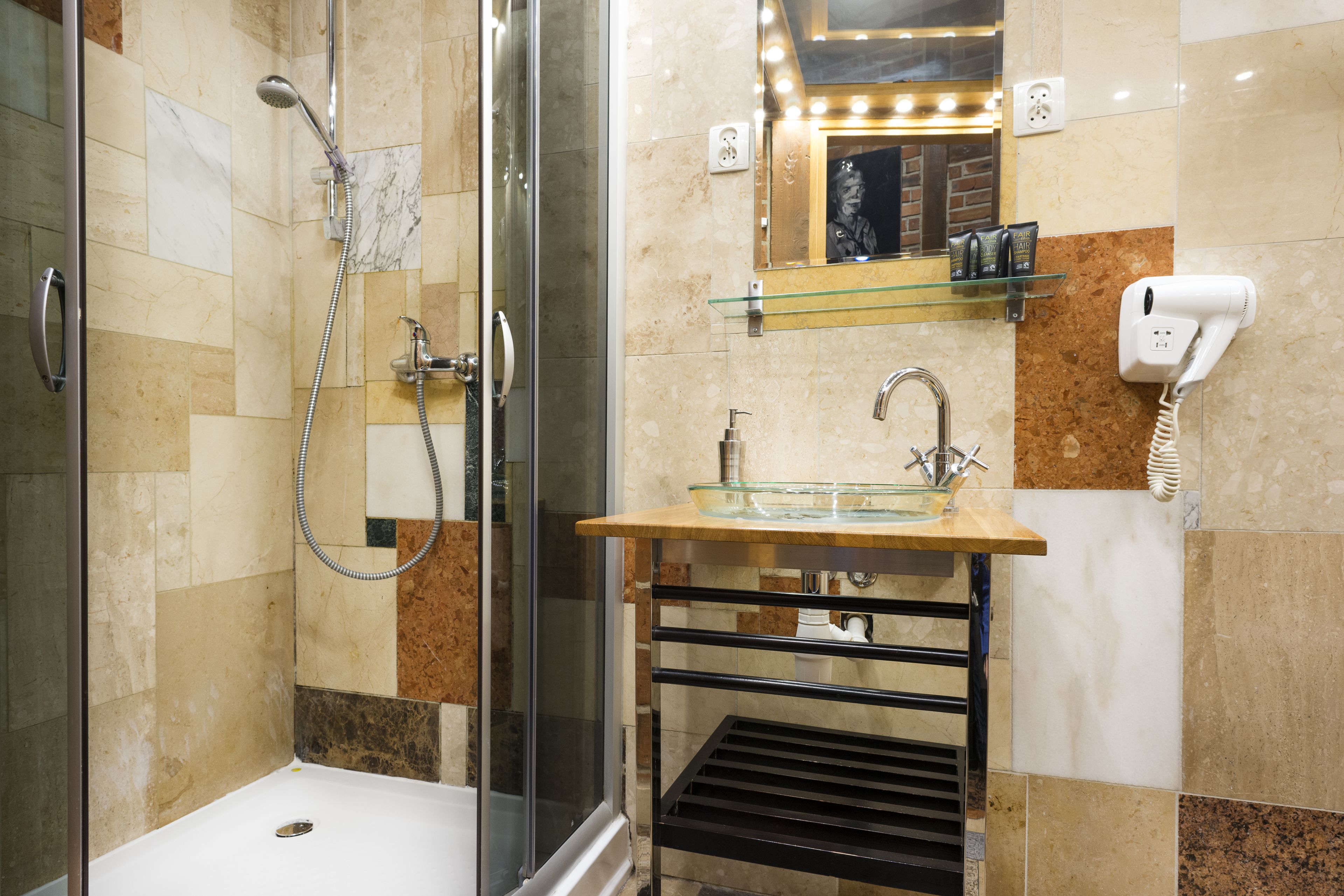 A modern bathroom with a glass shower enclosure, a glass sink with a marble countertop, a hairdryer on the wall, and a mirror above the sink.