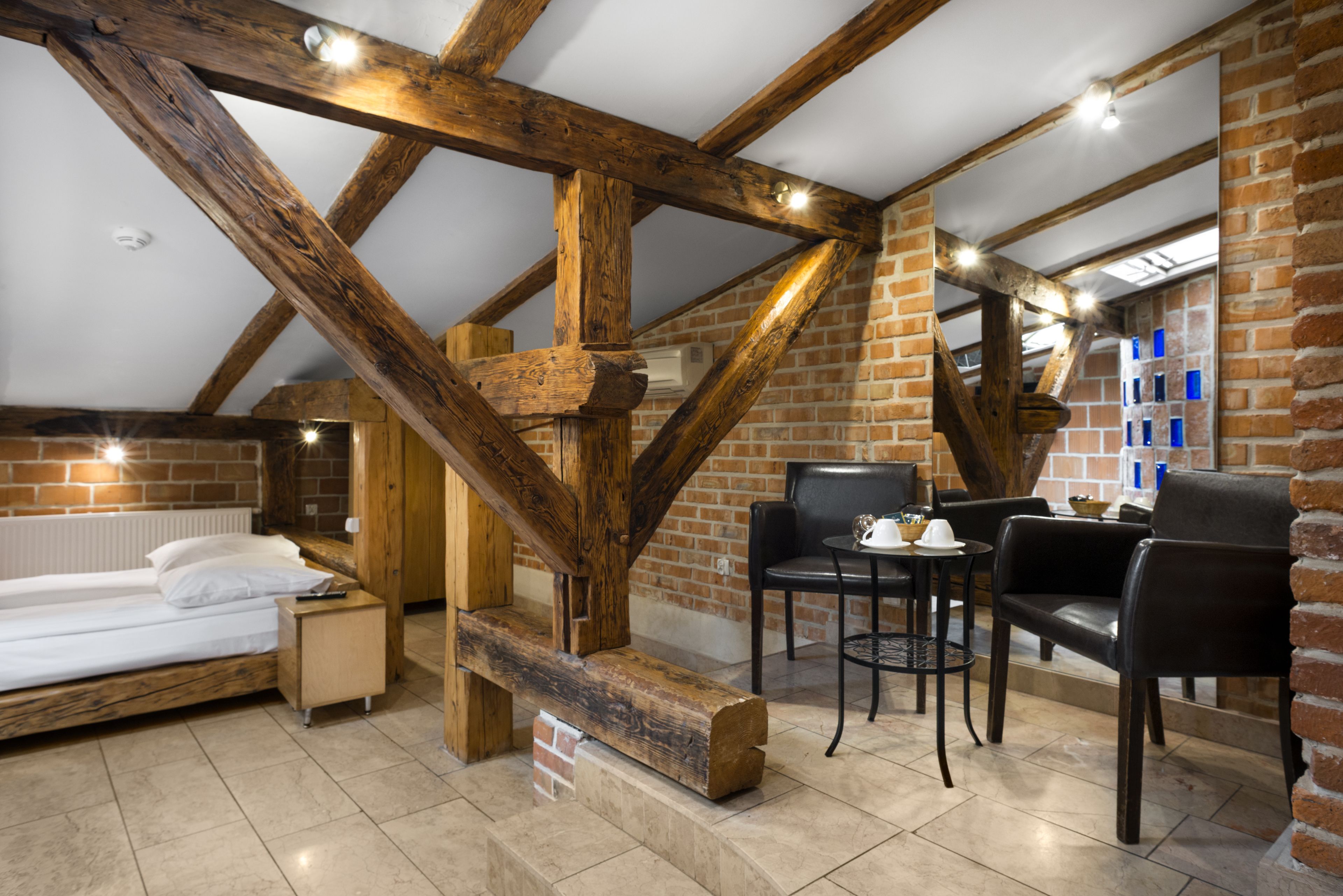 A bedroom with a wooden ceiling, a brick wall, a bed, a nightstand, and a small table with chairs.