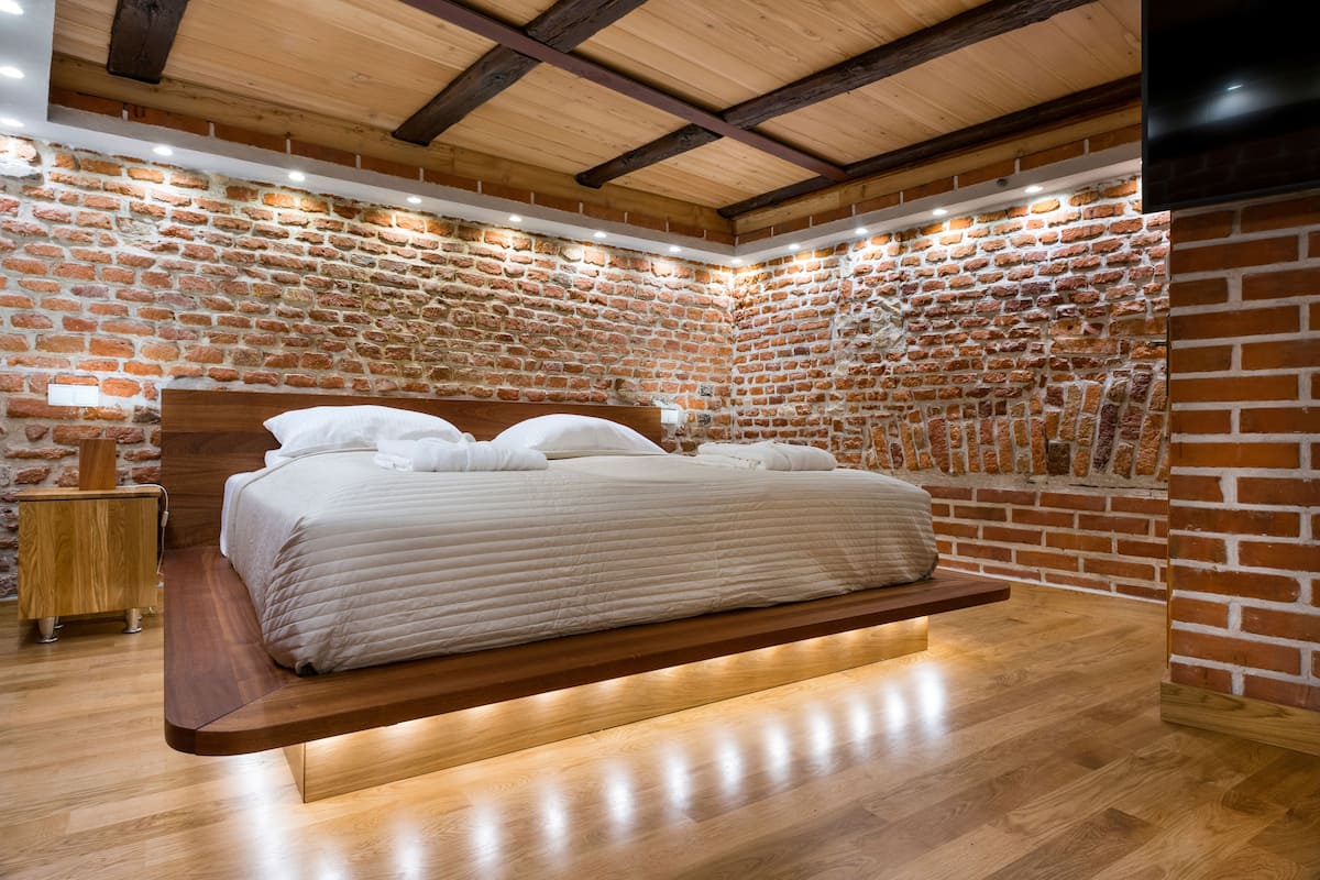 A bedroom with a brick wall, wooden ceiling beams, a bed with a wooden platform, and a bedside table.
