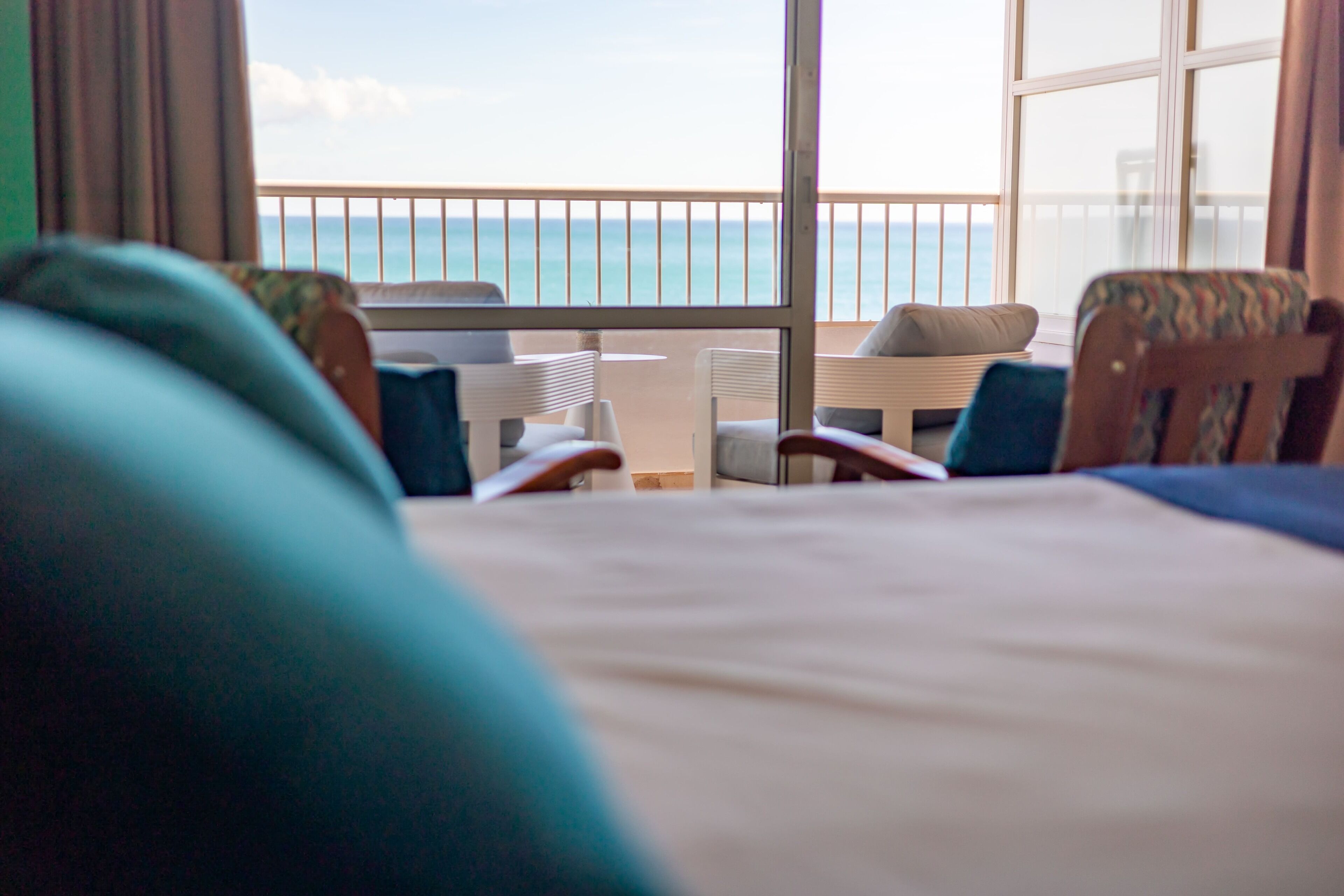 Een hotelkamer met een groot raam met uitzicht op zee, een bed en twee fauteuils met kussens.