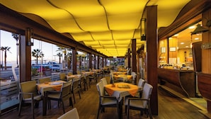 Un restaurante con suelo de madera, mesas con manteles anaranjados y vistas a palmeras y coches afuera.